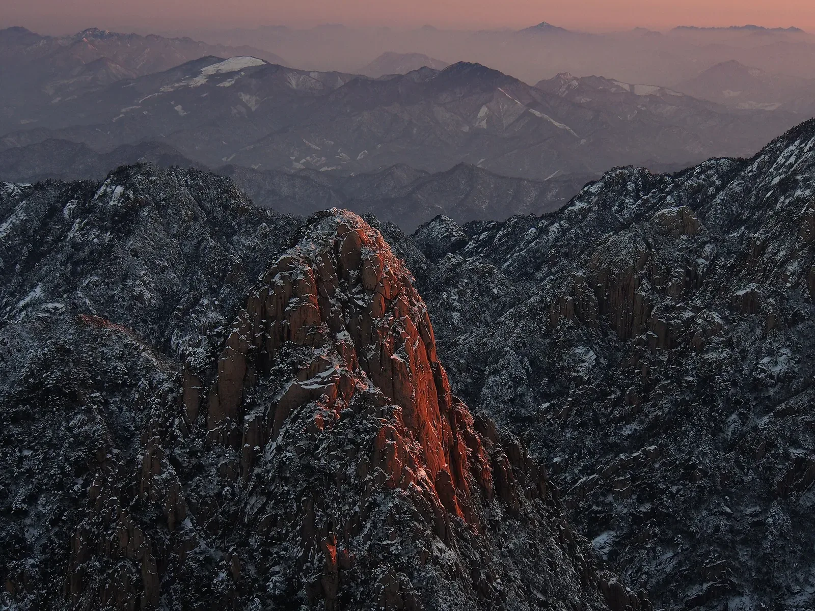 A snow-dusted granite peak in Huangshan at sunrise, lit by warm pink and orange first light, with the lower slopes and surrounding ranges in deep blue shadow and a pinkish-purple sky stretching to the horizon.