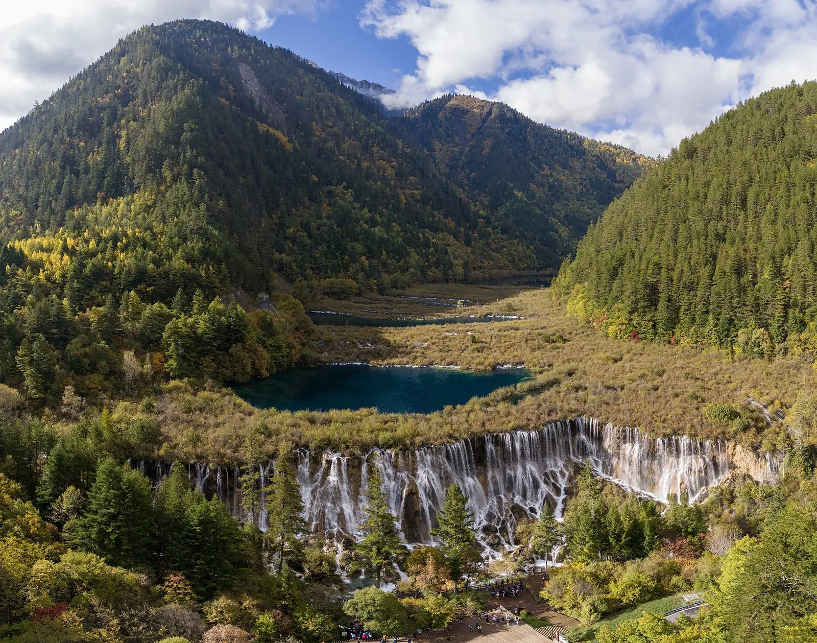 The Nuorilang Waterfall in Jiuzhaigou Valley, Sichuan — a wide travertine cascade pouring over a curved rim into a turquoise pool, framed by forested mountains with autumn gold and orange foliage, a long wooden boardwalk at the base, and a clear blue sky with white clouds above.