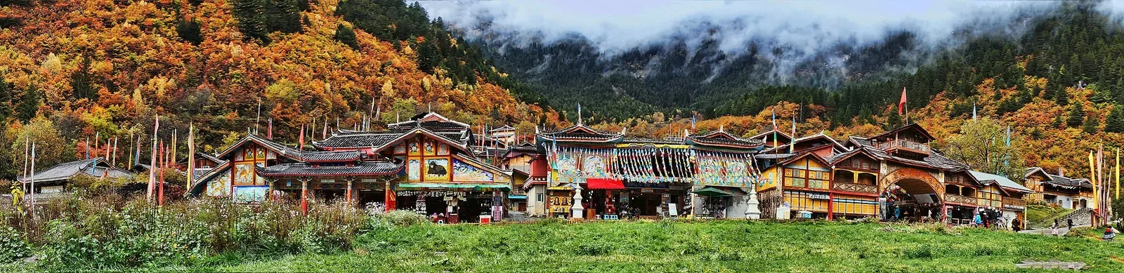 A wide panoramic view of Shuzheng Village in Jiuzhaigou Valley, Sichuan — a row of traditional Tibetan wooden houses with painted eaves, prayer flags, and stone walls along a green meadow, backed by forested mountains in brilliant autumn reds, oranges, and yellows, with wisps of cloud drifting through the peaks.