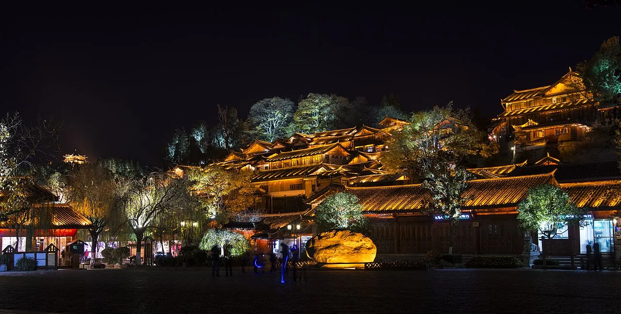 The old town of Lijiang at night, with a cascade of traditional Naxi wooden houses with tile roofs glowing in warm gold light up a hillside, and a few visitors walking in the cobblestone square below.
