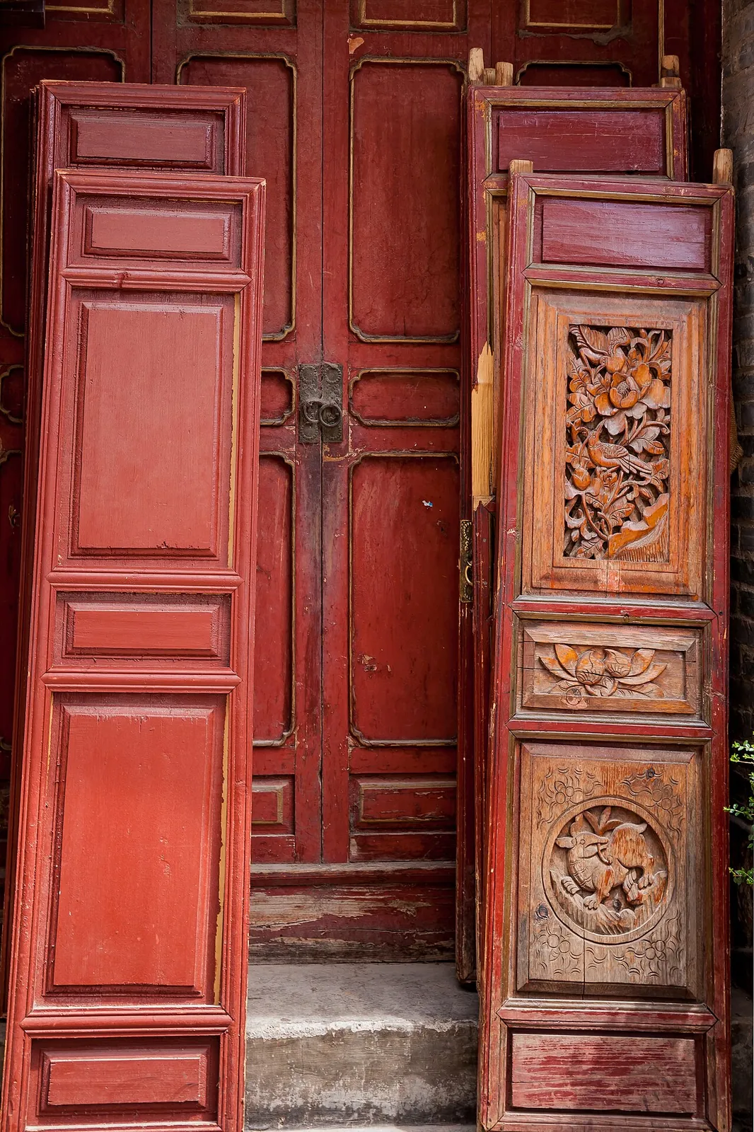Two ornate carved wooden doors in a courtyard in Lijiang's old town — one painted deep red with a brass lock, the other an aged natural-wood panel with intricate carvings of birds and flowers and a goat motif at the bottom.