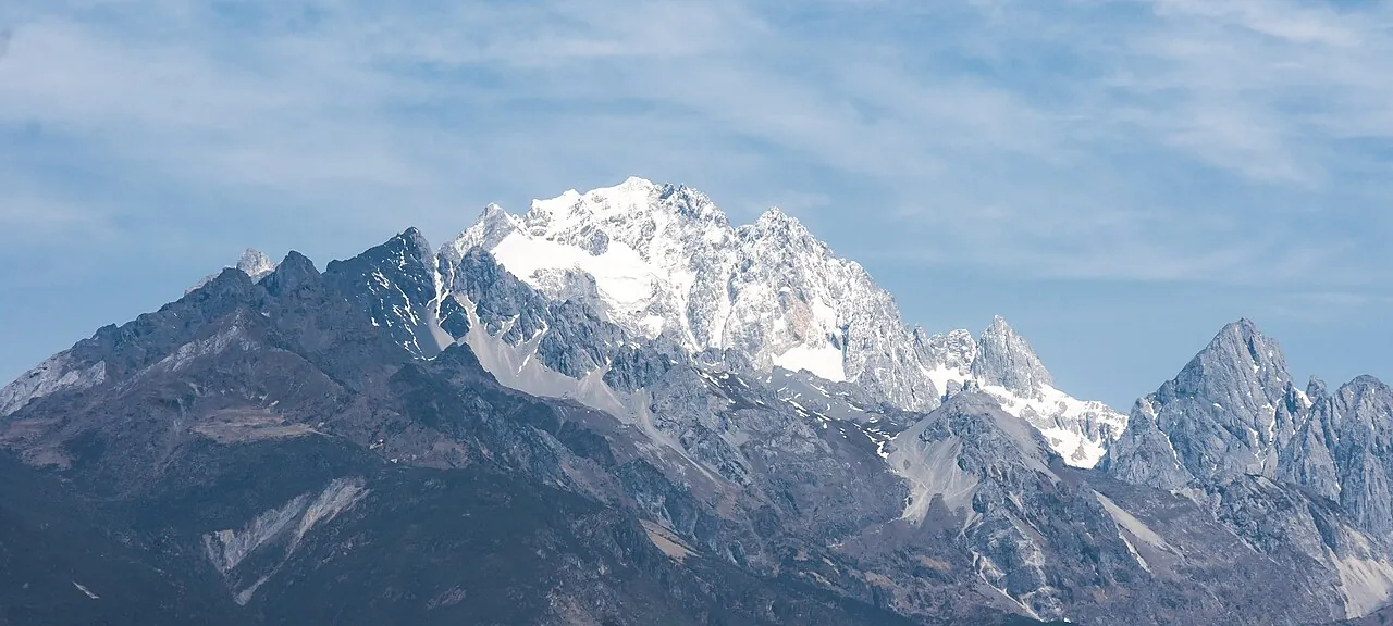 The snow-capped main peak of Jade Dragon Snow Mountain (Yulong Xueshan) near Lijiang, with jagged grey-black ridges below the white summit under a blue sky streaked with thin clouds.