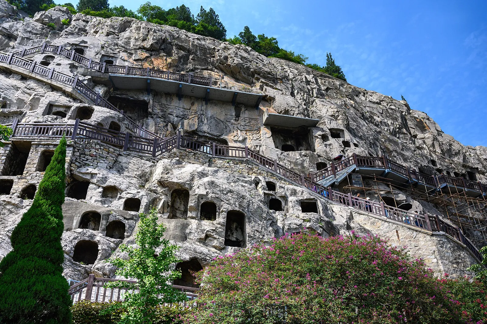 The Longmen Grottoes near Luoyang, a UNESCO World Heritage Site — a sheer limestone cliff face honeycombed with hundreds of Buddhist cave shrines and statuary carved between the 5th and 8th centuries, with wooden walkways climbing the cliff.
