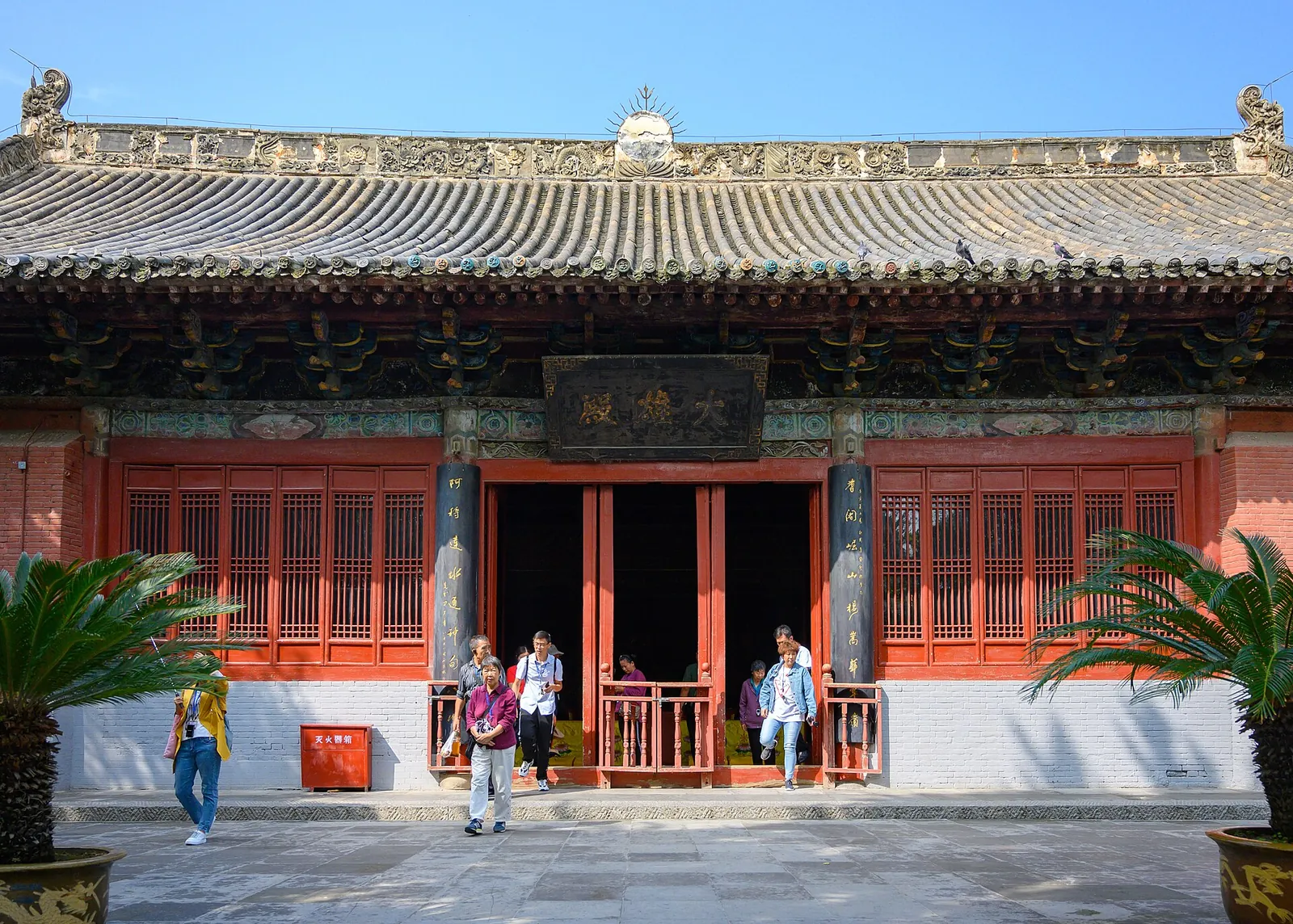 The entrance of the White Horse Temple in Luoyang — the first Buddhist temple in China, founded in 68 AD — with a red wooden facade, grey-tile roof, gold-painted eaves, and visitors walking out through the carved doors under a sunny sky.