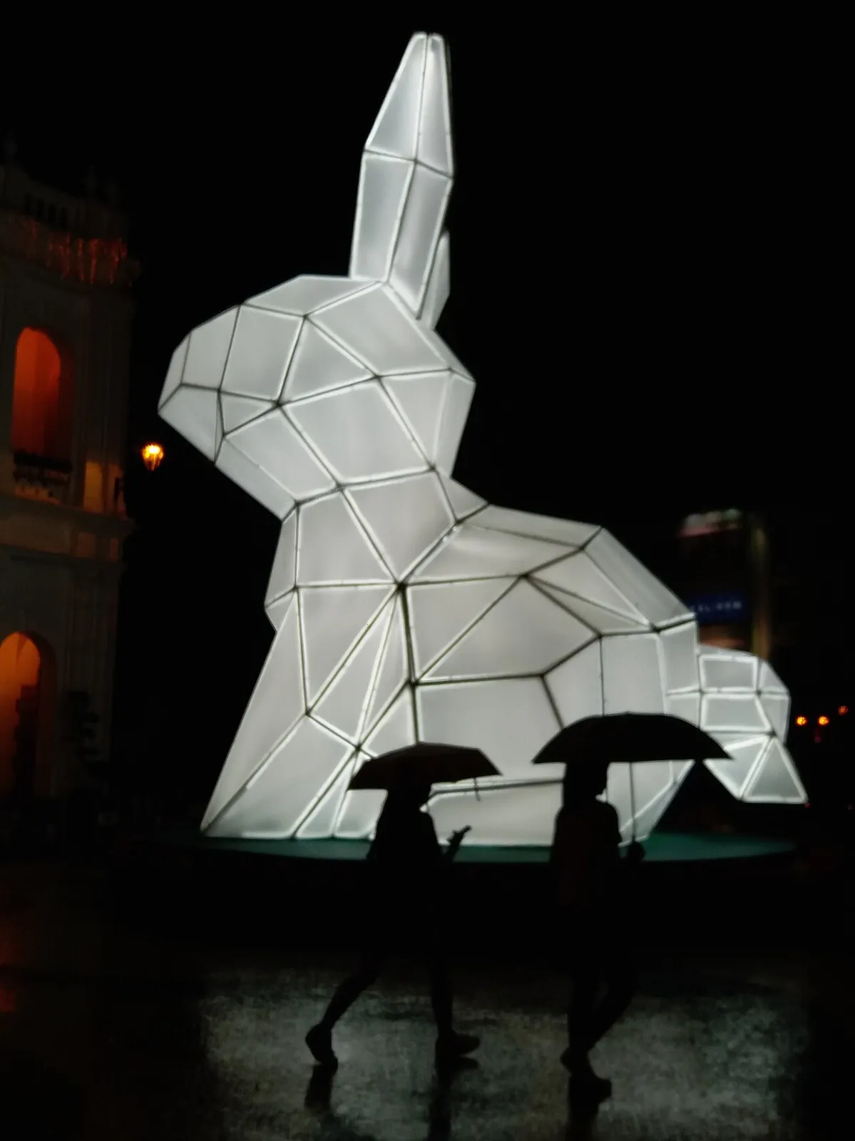 A large geometric rabbit lantern installation glowing white at night in Macau, with two people holding umbrellas walking past — part of a Lunar New Year light festival in the historic centre.
