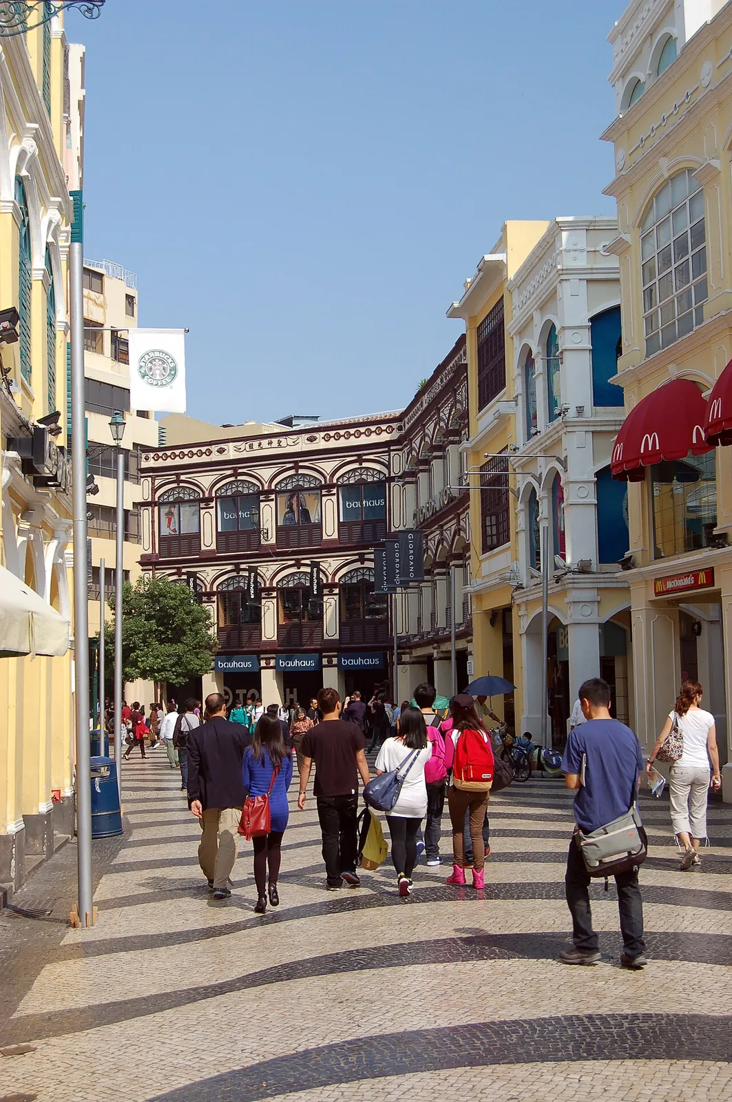 Senado Square in Macau, with tourists walking among the iconic pastel-yellow Portuguese colonial buildings, traditional shop signs, and a Bauhaus storefront on a sunny day.