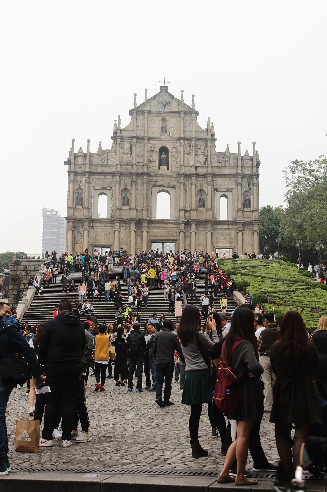 The full Baroque facade of the Ruins of St Paul's in Macau, with a massive crowd of visitors gathered on the stone steps and cobblestone plaza in front — the most iconic landmark of the city.