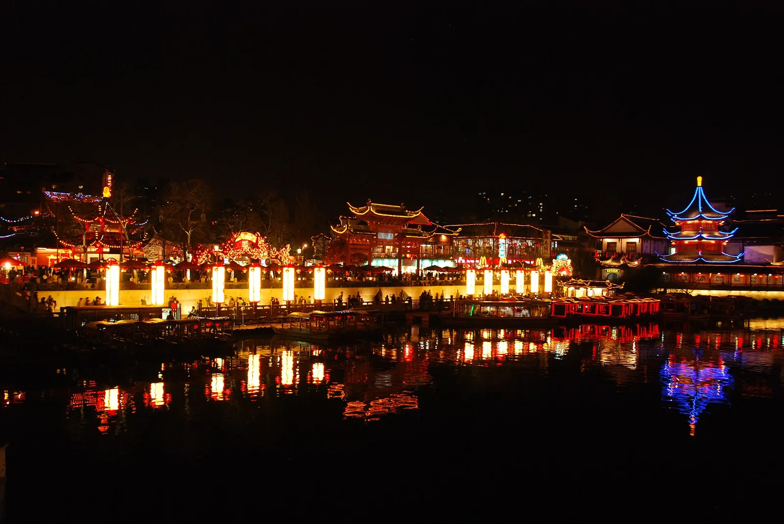 The Confucius Temple (Fuzimiao) and the Qinhuai River in Nanjing at night, with red and gold lanterns reflecting in the dark water, a crowd of visitors along the riverbank, and traditional Chinese pavilions with upturned eaves lit up in blue neon.
