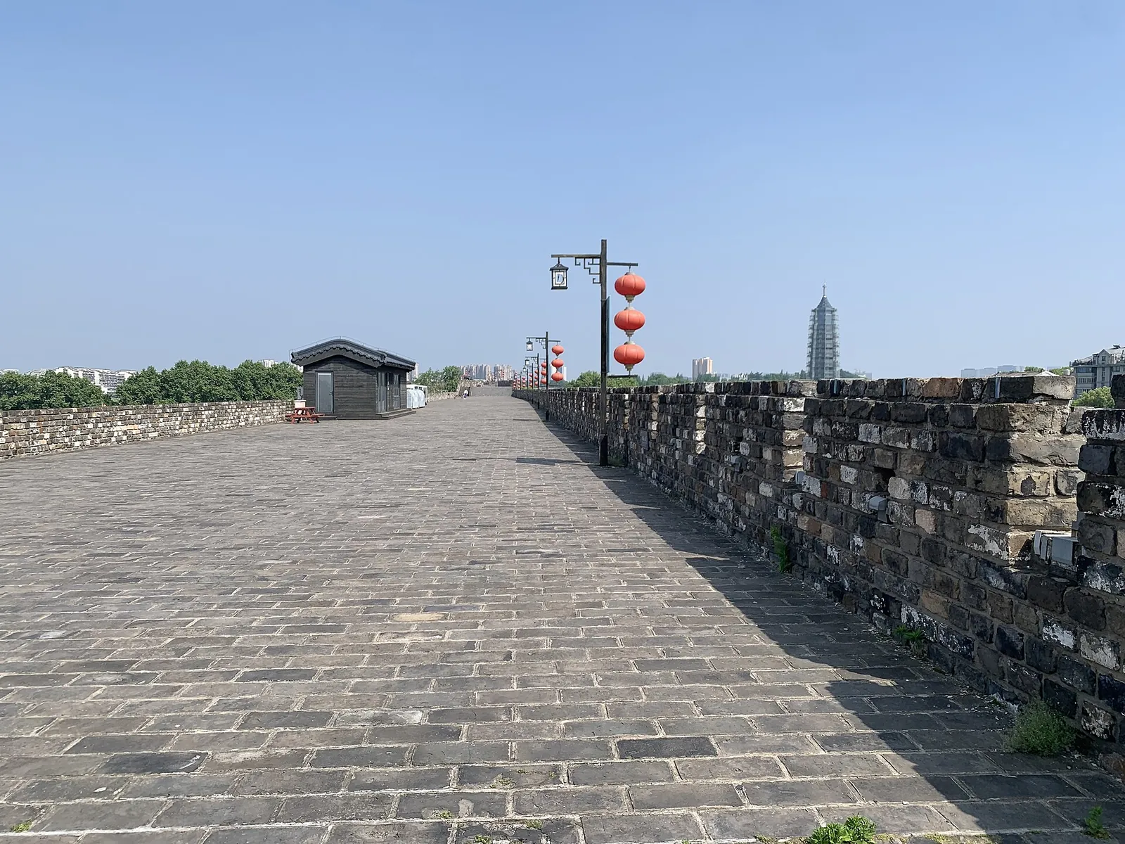 A wide stone-paved walkway on top of the ancient Nanjing City Wall (Ming Xiaoling), stretching into the distance with red lanterns lining the right side and a small wooden hut, with the modern Jinling pagoda of the Jiming Temple visible in the far distance.