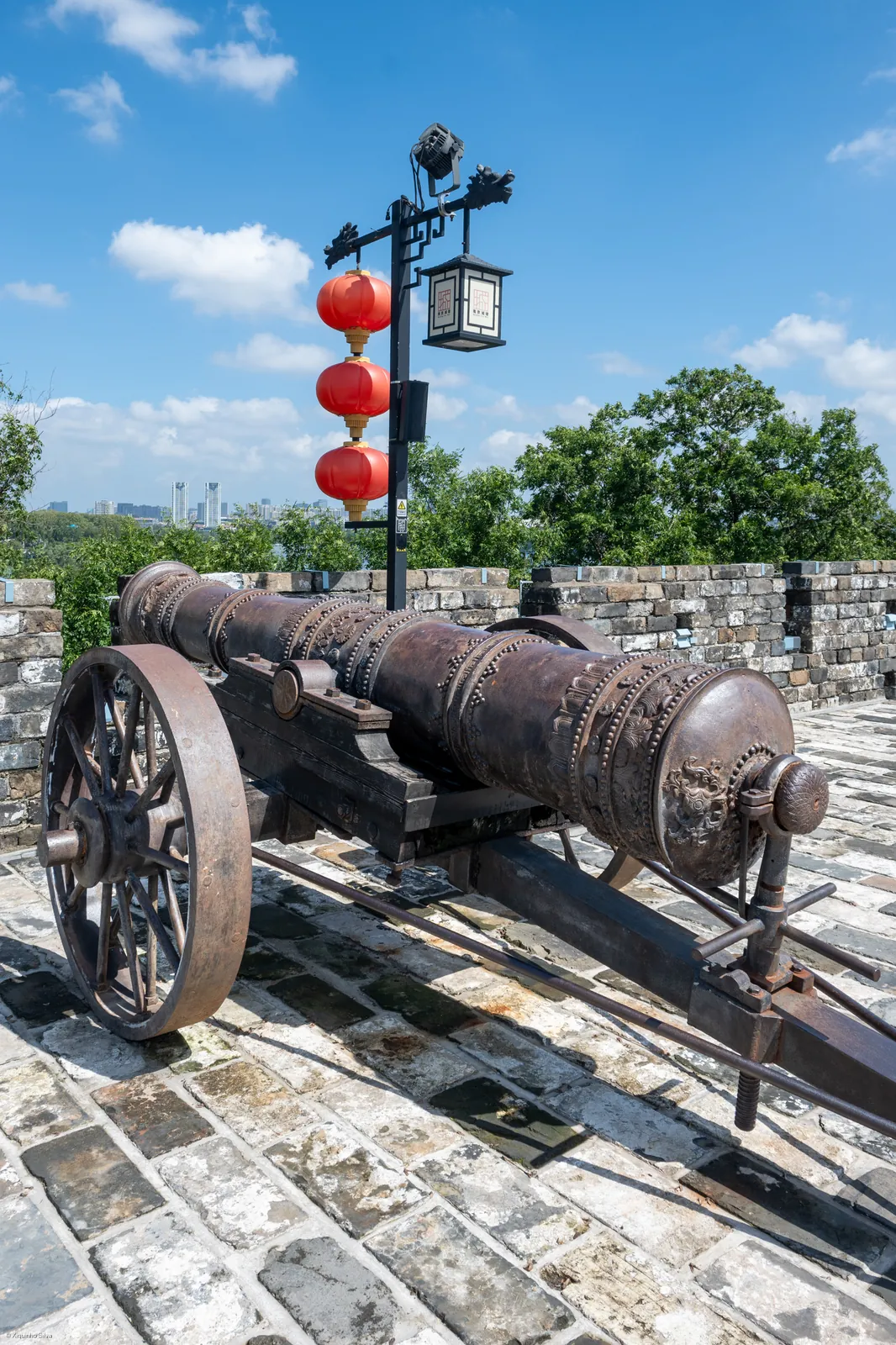 An old bronze cannon on the Nanjing City Wall, with elaborate dragon-and-cloud decorations along its barrel, parked on grey stone paving under red lanterns and a Chinese street sign, with the city skyline in the background.