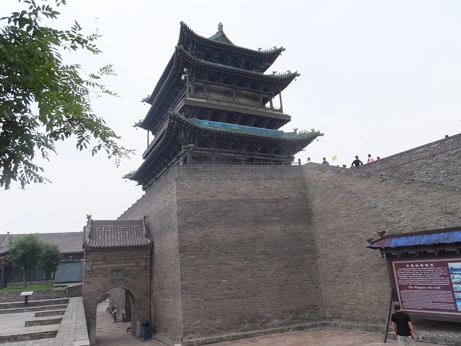 A corner watchtower of the ancient Pingyao city wall in Shanxi — a three-tiered wooden pagoda-style tower on top of massive grey brick battlements, with visitors walking on the wall and an information panel about the wall in the foreground.