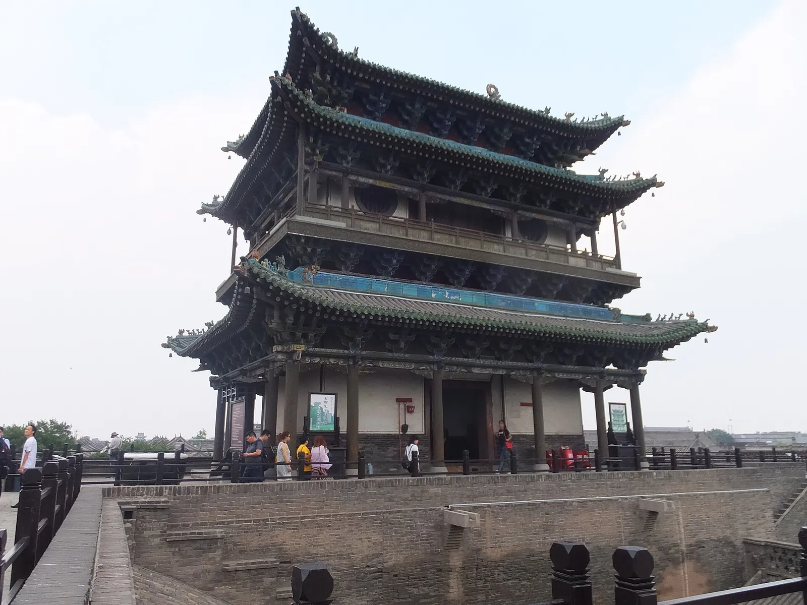 The North Gate of the Pingyao ancient city wall, with a tall multi-tiered wooden gate tower, a stone-paved ramp leading up to it, and several visitors walking up and down.
