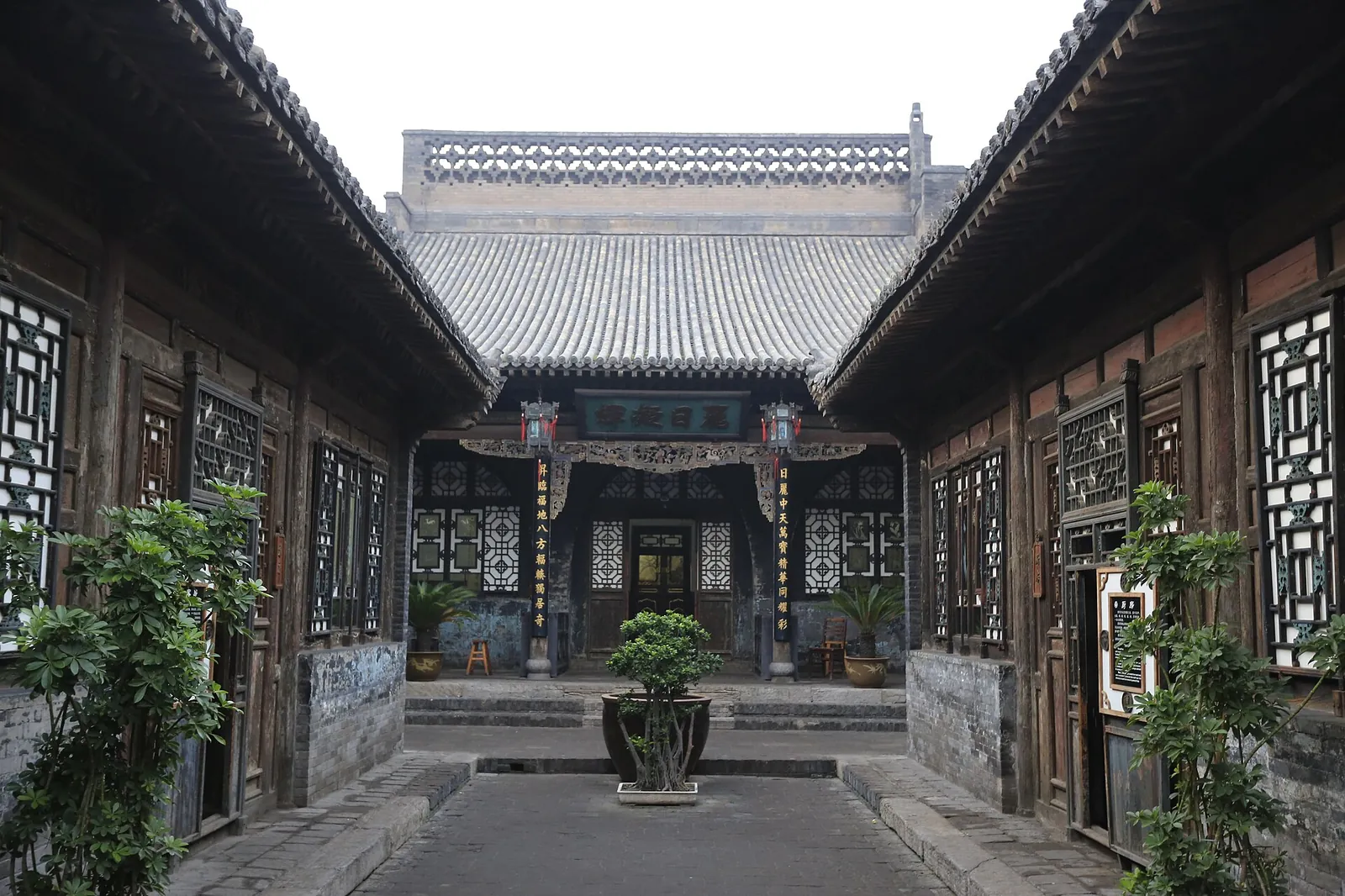 A traditional grey-tiled interior courtyard of the Rishengchang Draft Bank in Pingyao, China's first bank, with wooden lattice windows, stone steps, potted plants, and a deep wooden hall at the back.