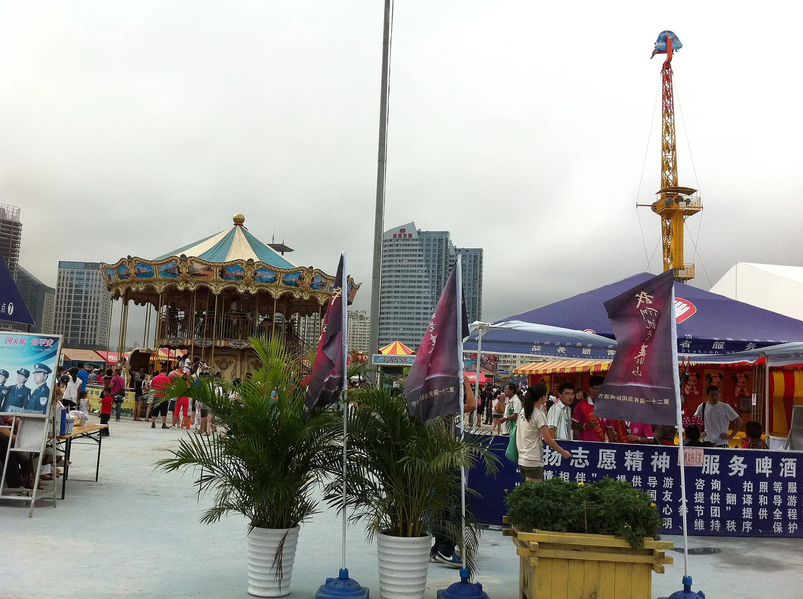 A vintage carousel and blue-tented beer booths at the Qingdao International Beer Festival, with hundreds of visitors strolling the seaside venue, the city's modern skyline rising behind.
