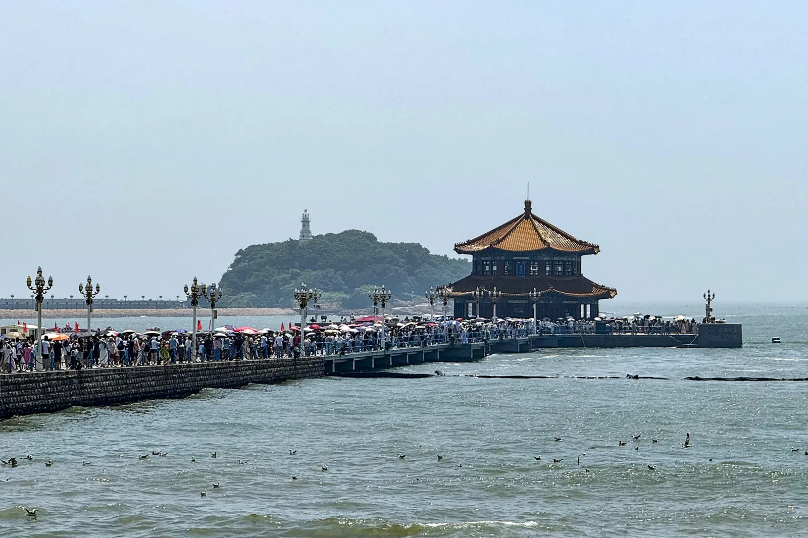 The Zhanqiao Pier in Qingdao, a 440-meter-long pier stretching into the yellow sea, crowded with visitors under umbrellas, with the octagonal Huilan Pavilion at the far end and Little Qingdao Isle and a white lighthouse in the background.