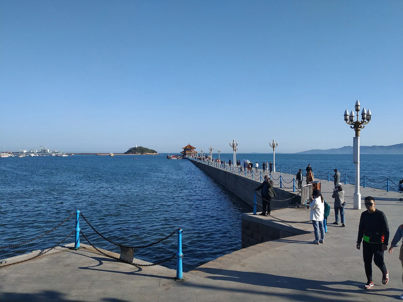 A wide view of Zhanqiao Pier in Qingdao on a clear day, with local walkers strolling along the granite walkway, the Huilan Pavilion at the far end and the Shandong coastline in the distance.