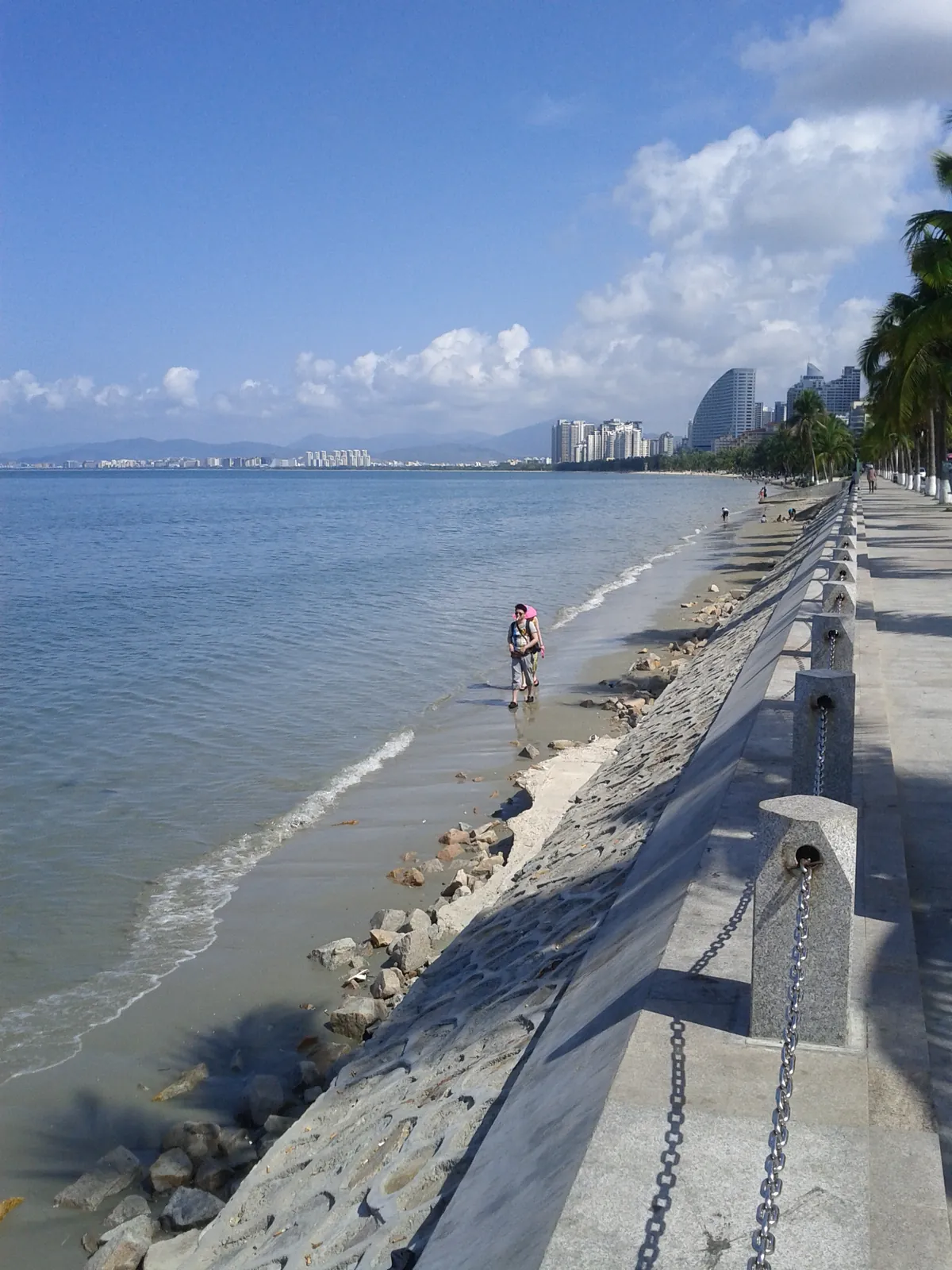 A coastal promenade in Sanya, Hainan, with palm trees, a stone sea wall, and a local woman walking on the sand by the South China Sea, with the city's modern skyline and mountains in the distance.