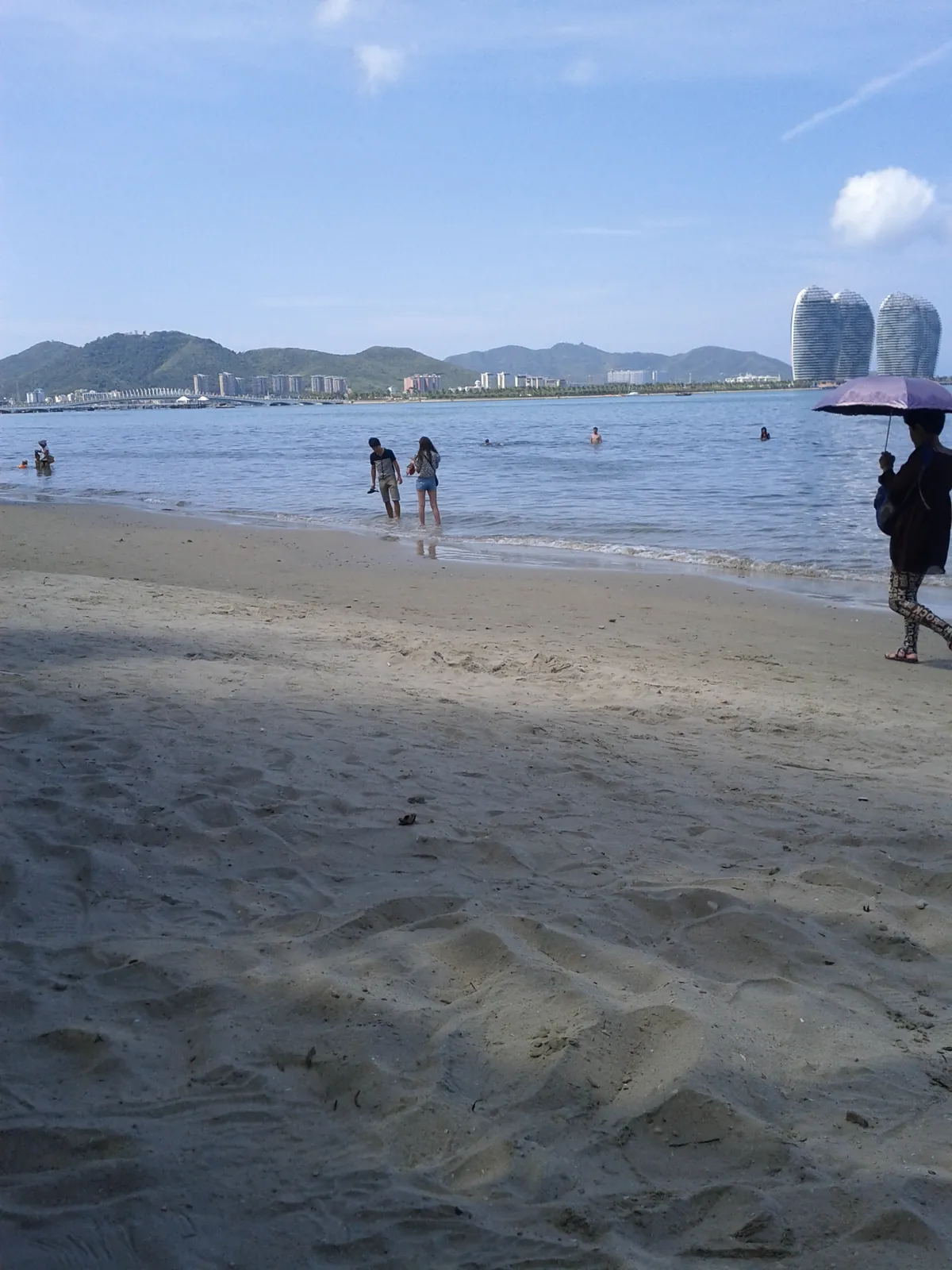A clean white-sand beach in Sanya on a clear day, with people wading in the warm water and Phoenix Island's curved white towers visible across the bay.