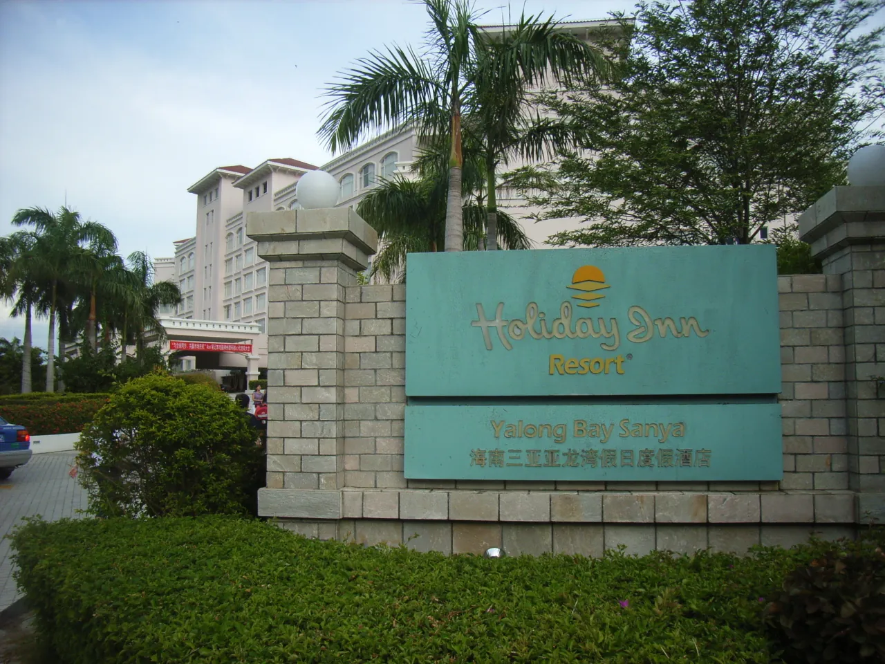 The Holiday Inn Resort entrance at Yalong Bay, Sanya — a teal and yellow signage on a stone base with tropical palm trees and the resort's pink multi-storey building behind.
