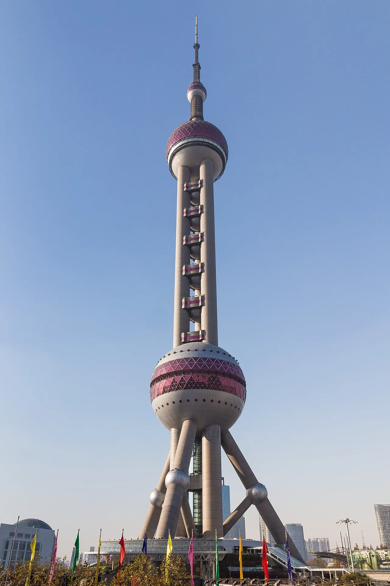The Oriental Pearl Tower in Shanghai's Pudong district on a clear day — the 468m futuristic TV tower with its distinctive pink-and-silver spheres and observation decks, the lower city skyline in the background and colored pennant flags fluttering in the foreground at the base of the tower, the most iconic landmark of the Shanghai skyline completed in 1994