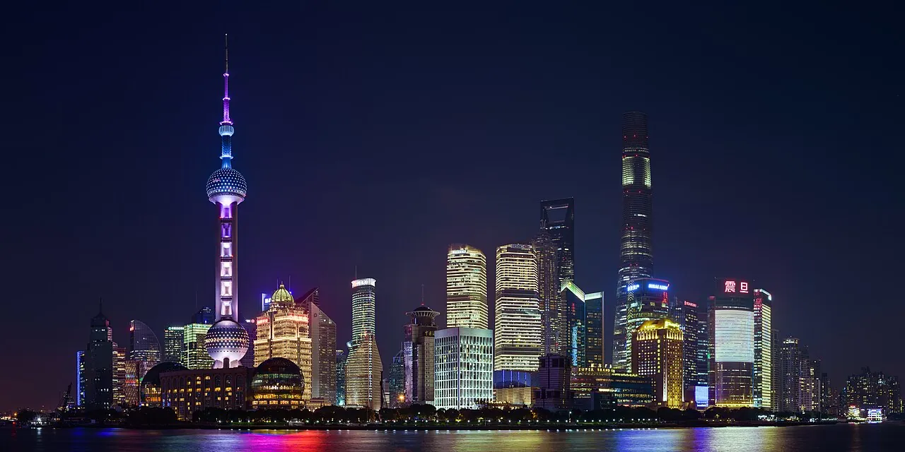 The Pudong skyline at night seen from the Bund across the Huangpu River — the Oriental Pearl Tower lit in purple on the left, the Shanghai Tower (632m, tallest in China) on the right, and the IFC, Jin Mao, and Shanghai World Financial Center clustered in between, the lights reflected in the calm water of the river in the foreground, the most-cited panoramic view of Shanghai's modern skyline