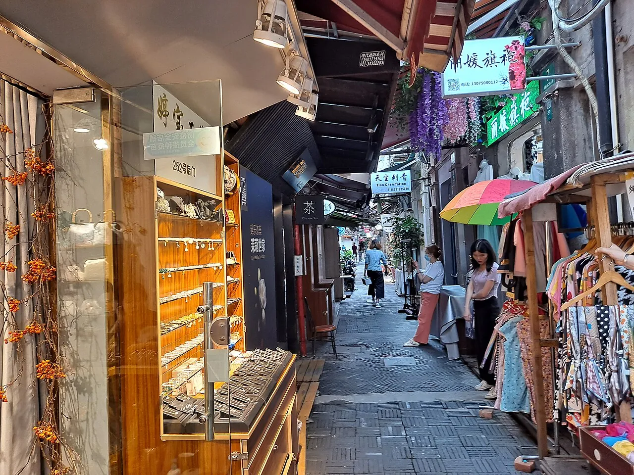A narrow lane in the Tianzifang arts district in Shanghai's former French Concession — converted 1920s shikumen stone-gate houses lining both sides of the stone-paved alley, small boutiques, tea shops and craft stores with signs in Chinese, a young woman in a white t-shirt looking at her phone in the foreground and a few other visitors walking the lane further down, the most atmospheric shopping and cafe district in central Shanghai
