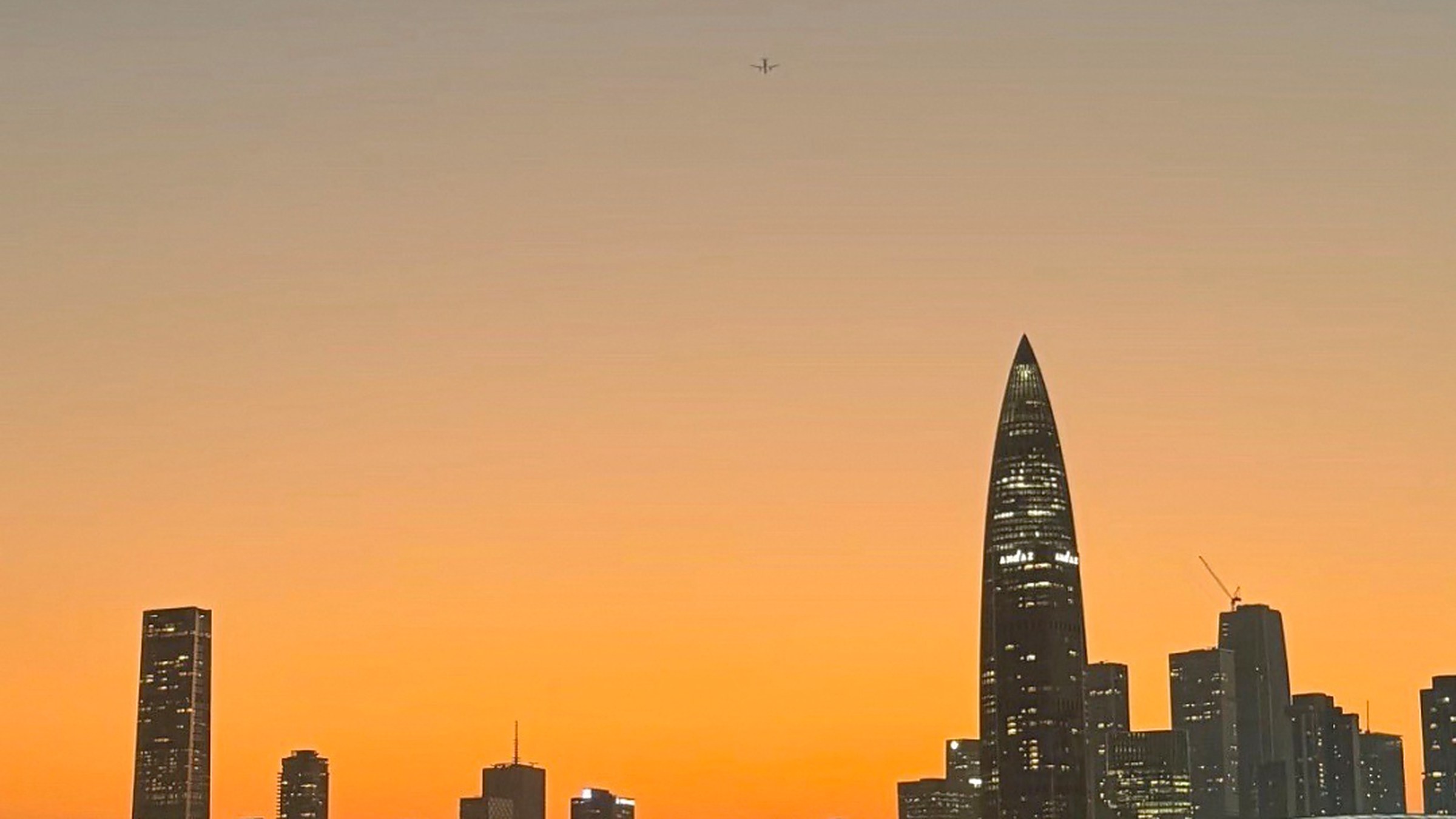Shenzhen Bay Park at sunset — the Spring Bamboo tower (Spring Bamboo shoot skyscraper) silhouetted against an orange and pink sky, with the bay-side promenade in the foreground