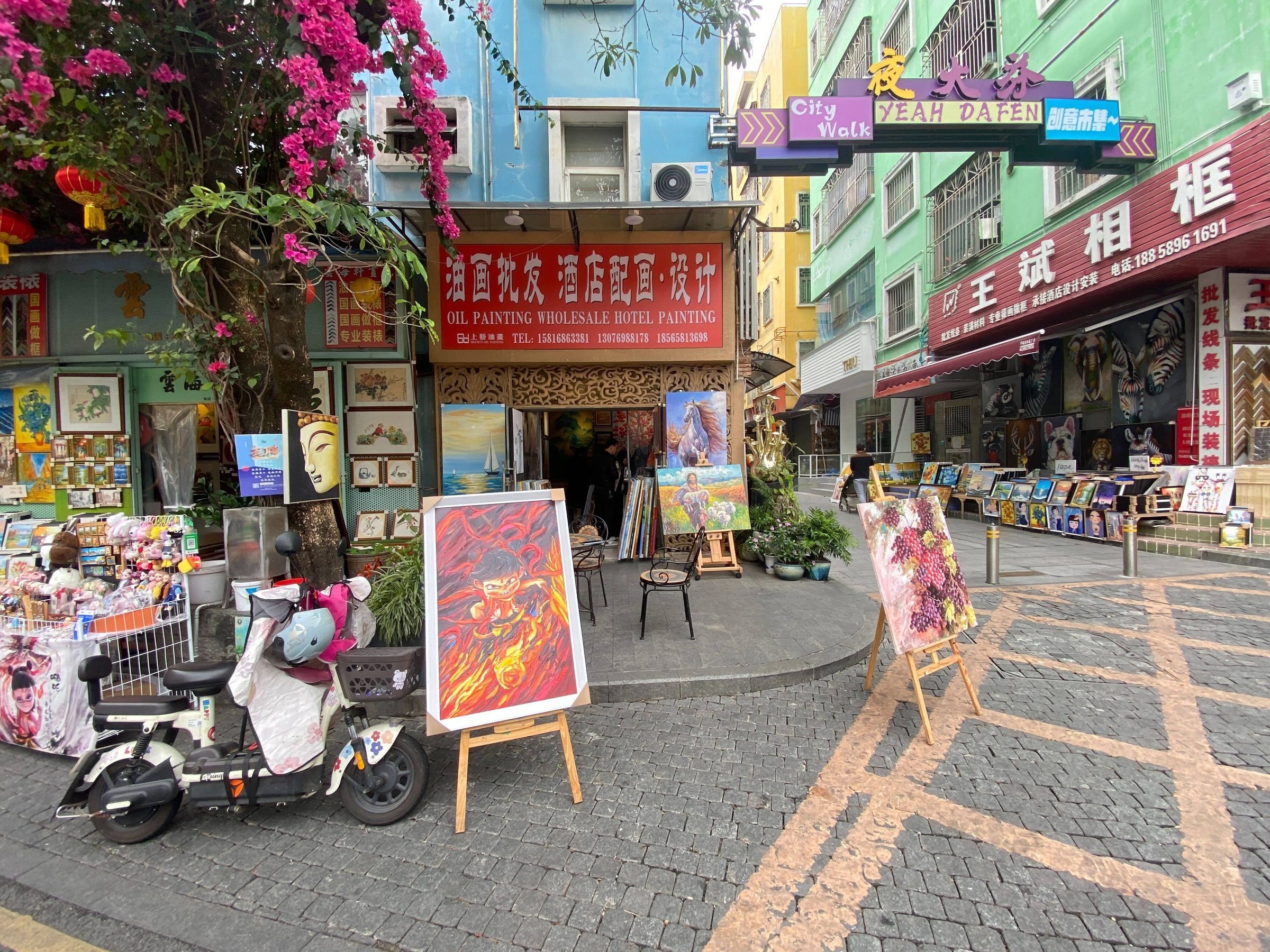 Dafen Oil Painting Village — paintings displayed on easels outside studios along the village street, with artists working in the doorways