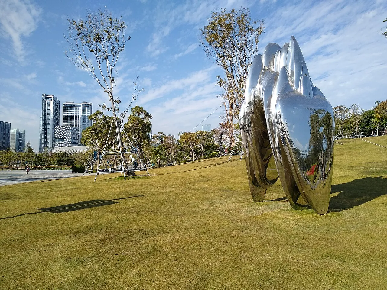 A polished stainless-steel hand sculpture by the sea in Shenzhen Bay Park, with a wide green lawn and the Houhai high-rise skyline in the background.