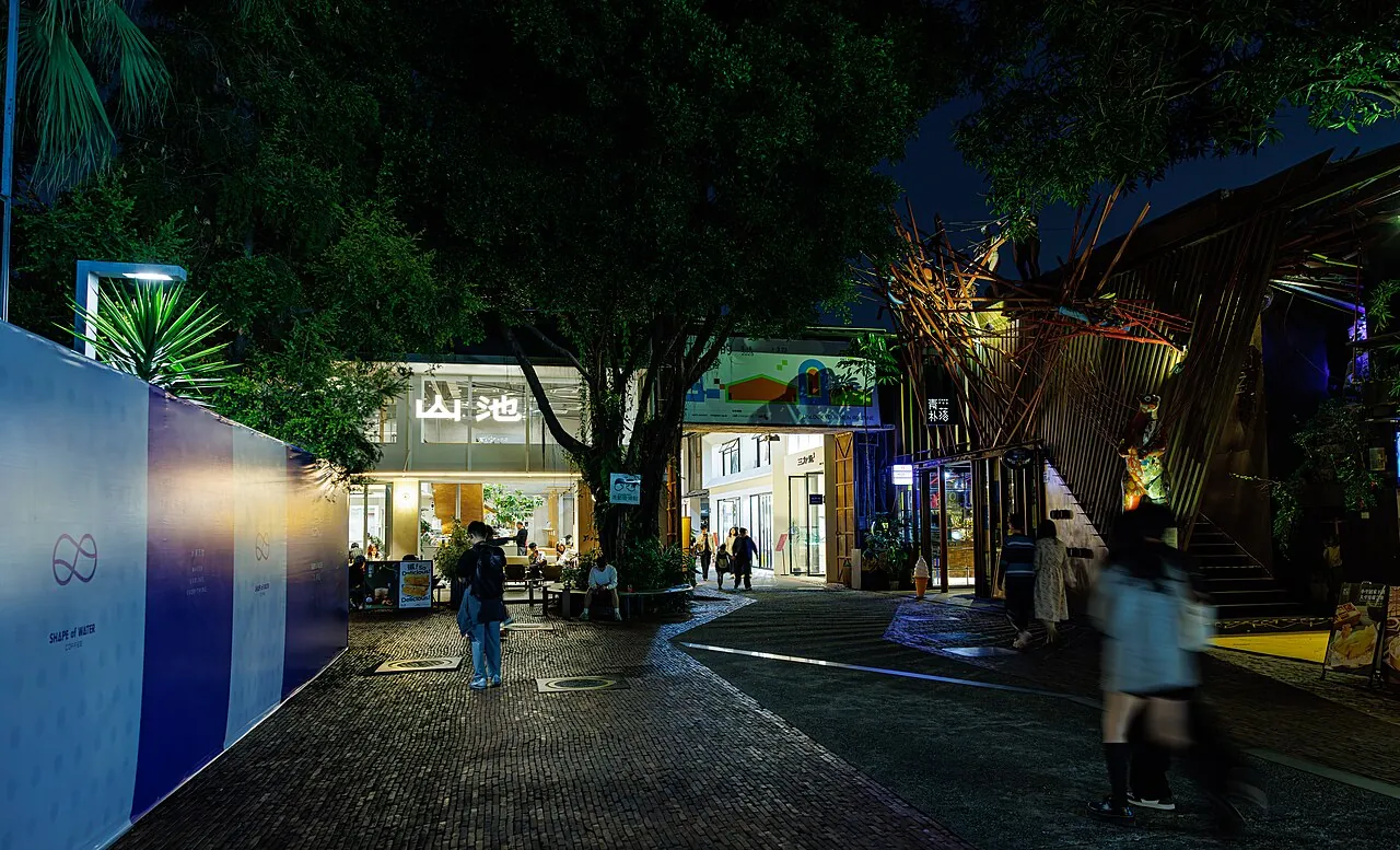 Visitors strolling through the OCT Loft creative district in Shenzhen at night, with a tree-lit café and a bamboo-wrapped architectural structure glowing along the cobblestone street.