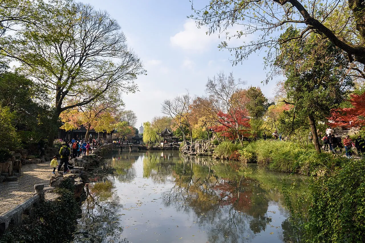 The Humble Administrator's Garden in Suzhou in autumn, with a still pond reflecting pavilions, willows, and a fiery canopy of red and orange maple leaves, with visitors walking along the covered walkway.