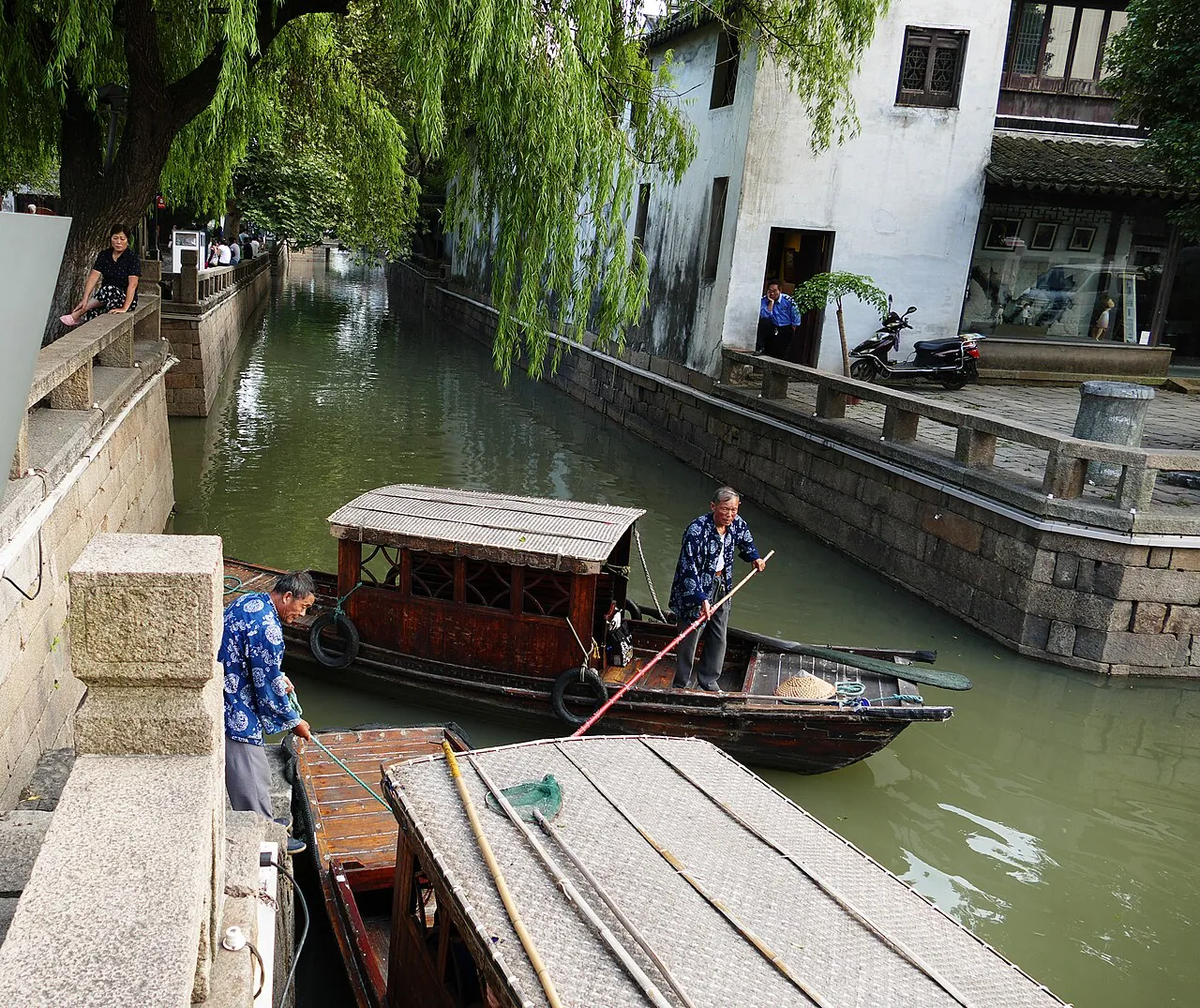 A narrow Pingjiang canal in Suzhou's old town, with two elderly men in blue shirts working a wooden boat under willow trees, whitewashed houses lining the water and an e-scooter parked on the stone embankment.