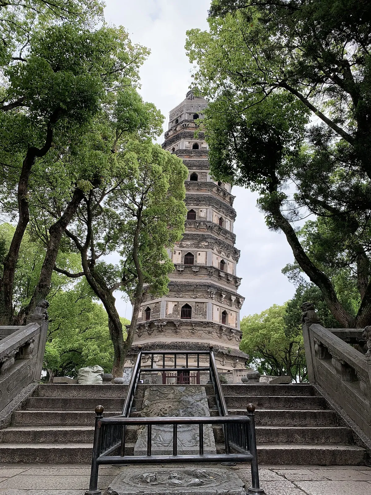 The leaning Tiger Hill Pagoda (Yunyan Si) in Suzhou, a seven-storey octagonal brick tower visibly tilted to one side, framed by lush trees and approached up a stone stairway with carved dragon panels.