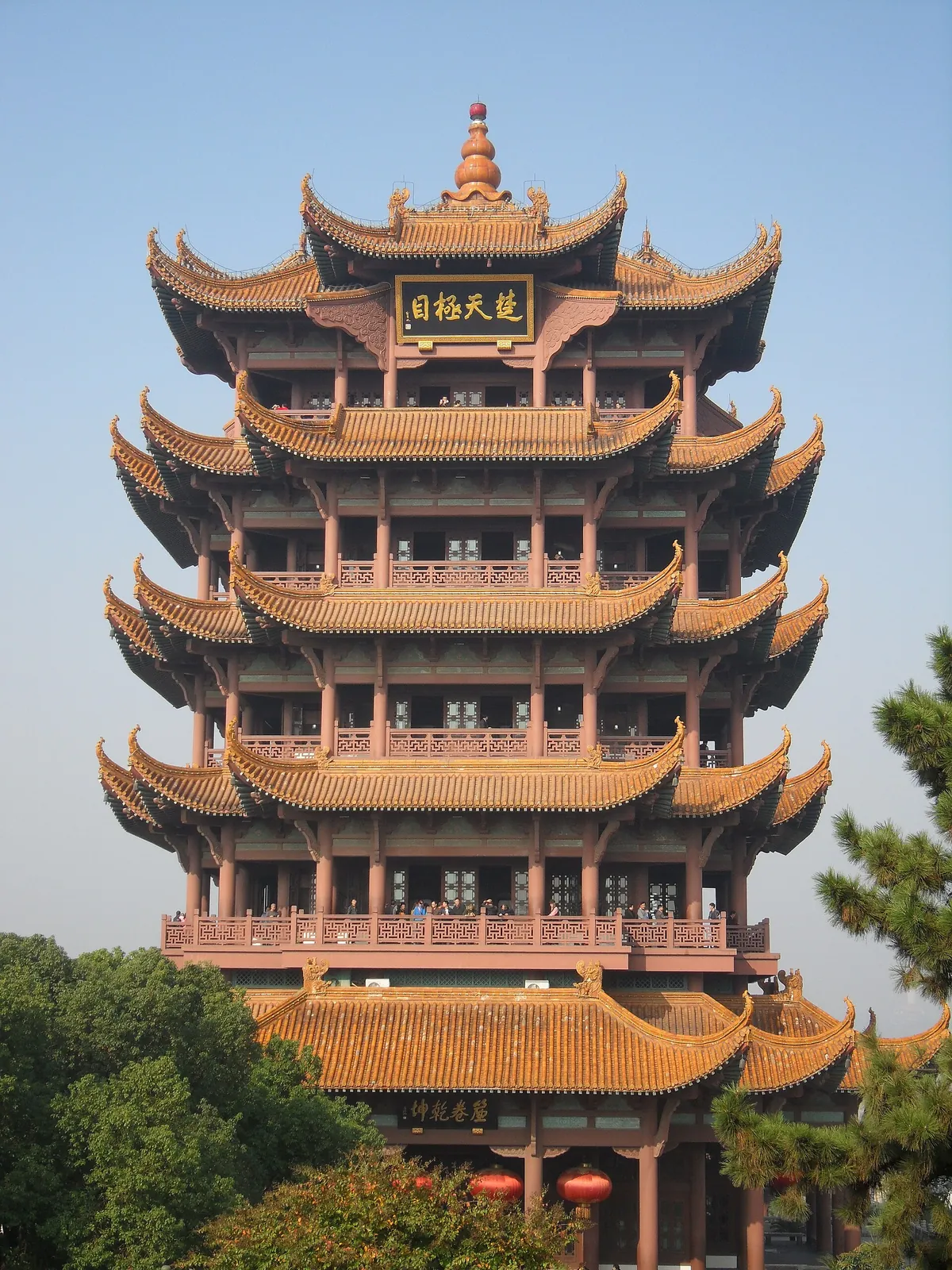 The Yellow Crane Tower (Huanghelou) in Wuhan — a five-tiered traditional pagoda with upturned eaves, golden tile roofs, a pumpkin-shaped finial, and visitors on the second-floor balcony, with pine trees and a clear blue sky in the background.