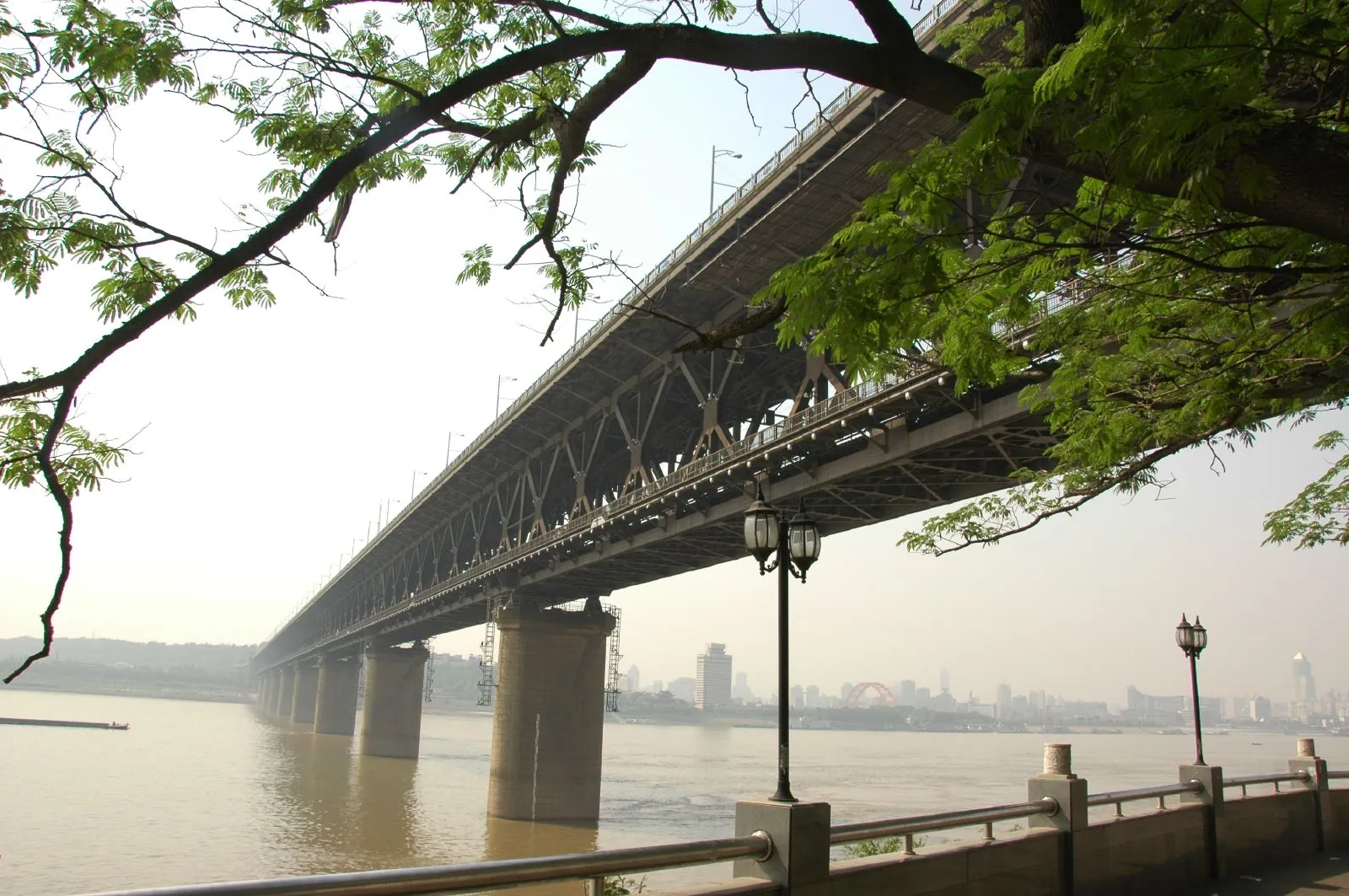 The Wuhan Yangtze River Bridge from below, showing the massive steel-truss underside of the road deck and a traditional Chinese pavilion perched on the rocky bank by the river.