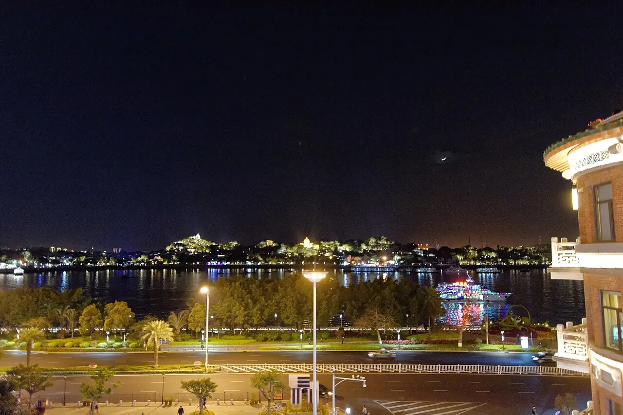 The night view of Gulangyu Island in Xiamen seen from the Zhongshan Road waterfront, with a string of golden-lit historic buildings on the island across the water and a few boats on the calm harbour.