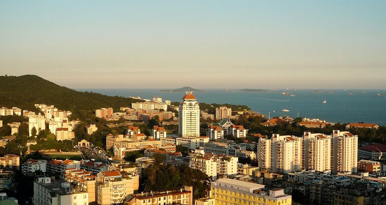A panoramic view of the Xiamen University campus at sunset, with the iconic twin white towers of the Jiageng building, lush green trees, and the South China Sea stretching to the horizon with cargo ships on the water.