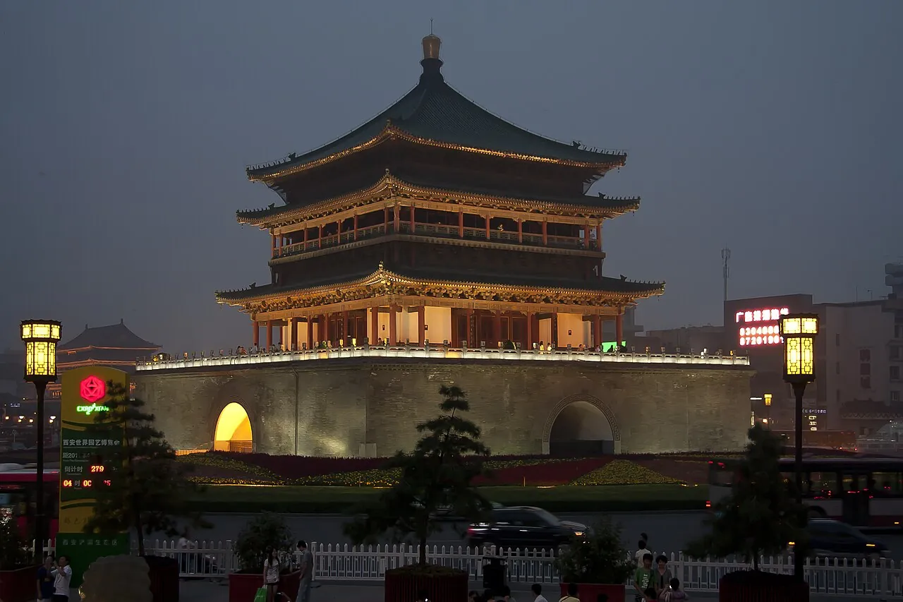 The Bell Tower of Xi'an, lit up at dusk, with visitors walking around its base in the busy city-center roundabout.