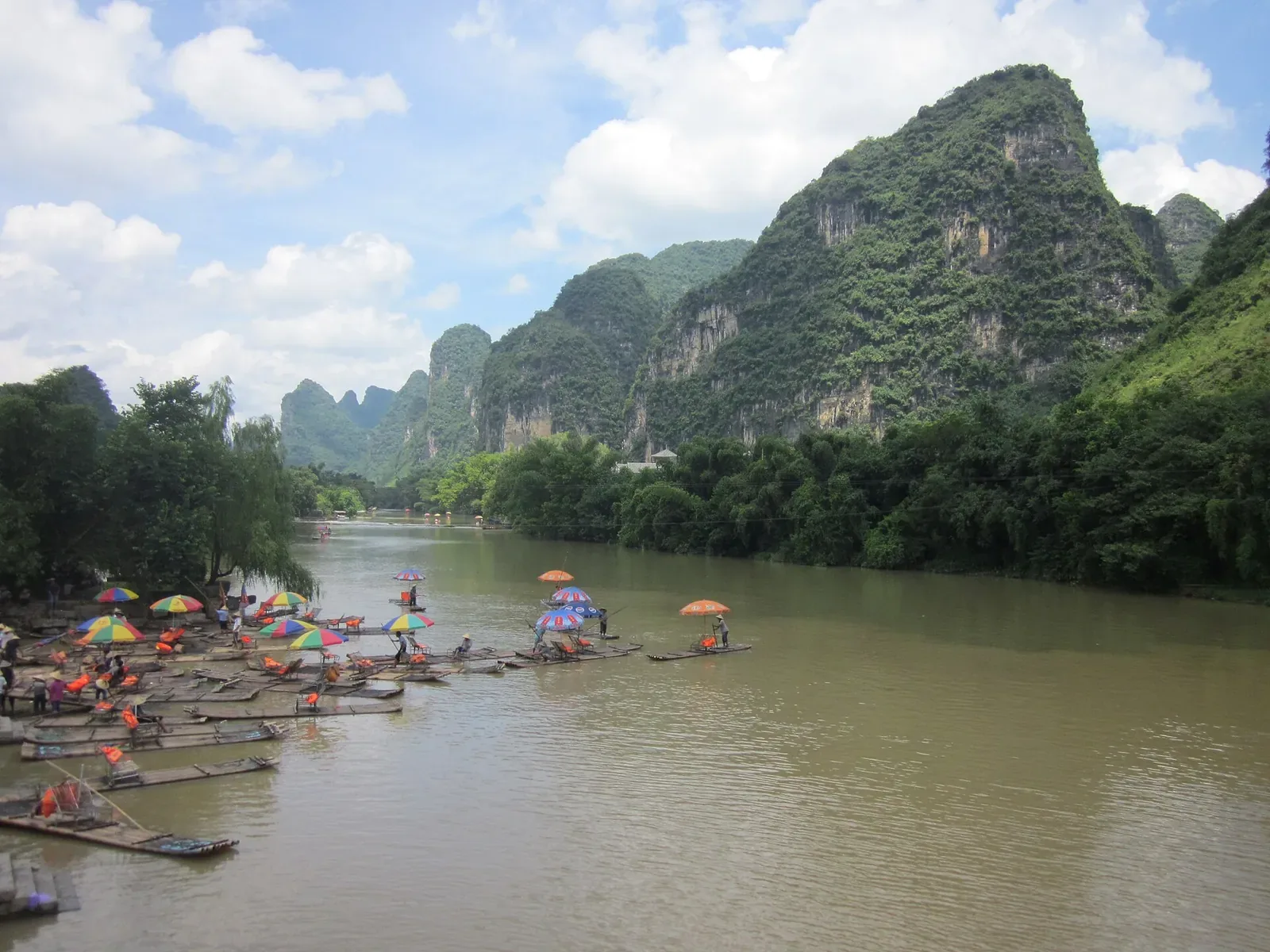 Bamboo rafts with colorful umbrellas floating on the Li River in Yangshuo, Guilin — dozens of traditional bamboo rafts clustered along the green riverbank, some already out on the calm jade-green water with bright red, blue, and yellow umbrellas shielding the raft operators, framed by dramatic cone-shaped karst limestone peaks rising under a partly cloudy blue sky.