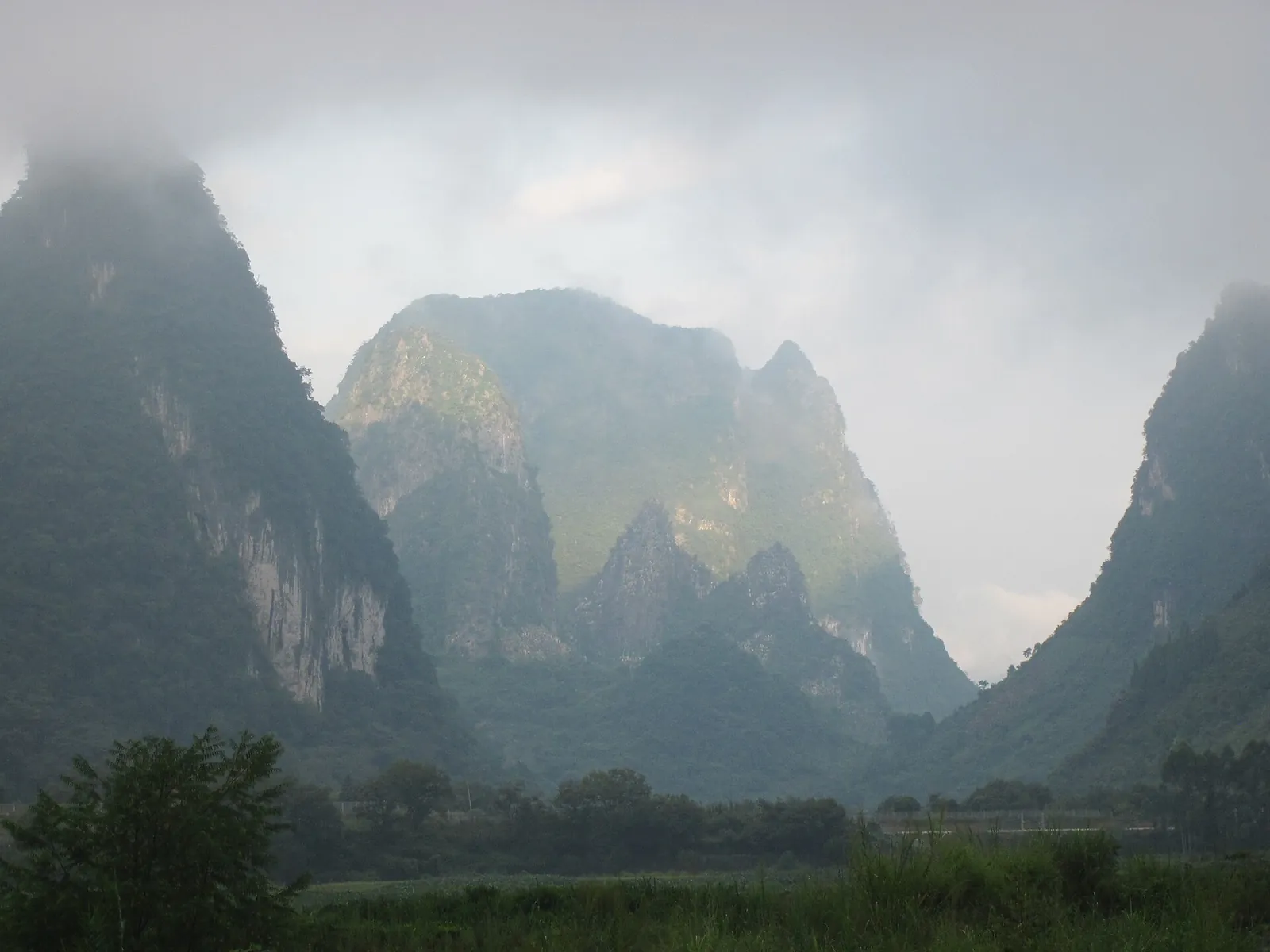 A misty view of the karst peaks of Yangshuo, Guilin — sharp cone-shaped limestone mountains rising dramatically from a green valley floor, partially shrouded in low-lying cloud and mist under an overcast grey sky, with grass and trees in the foreground.