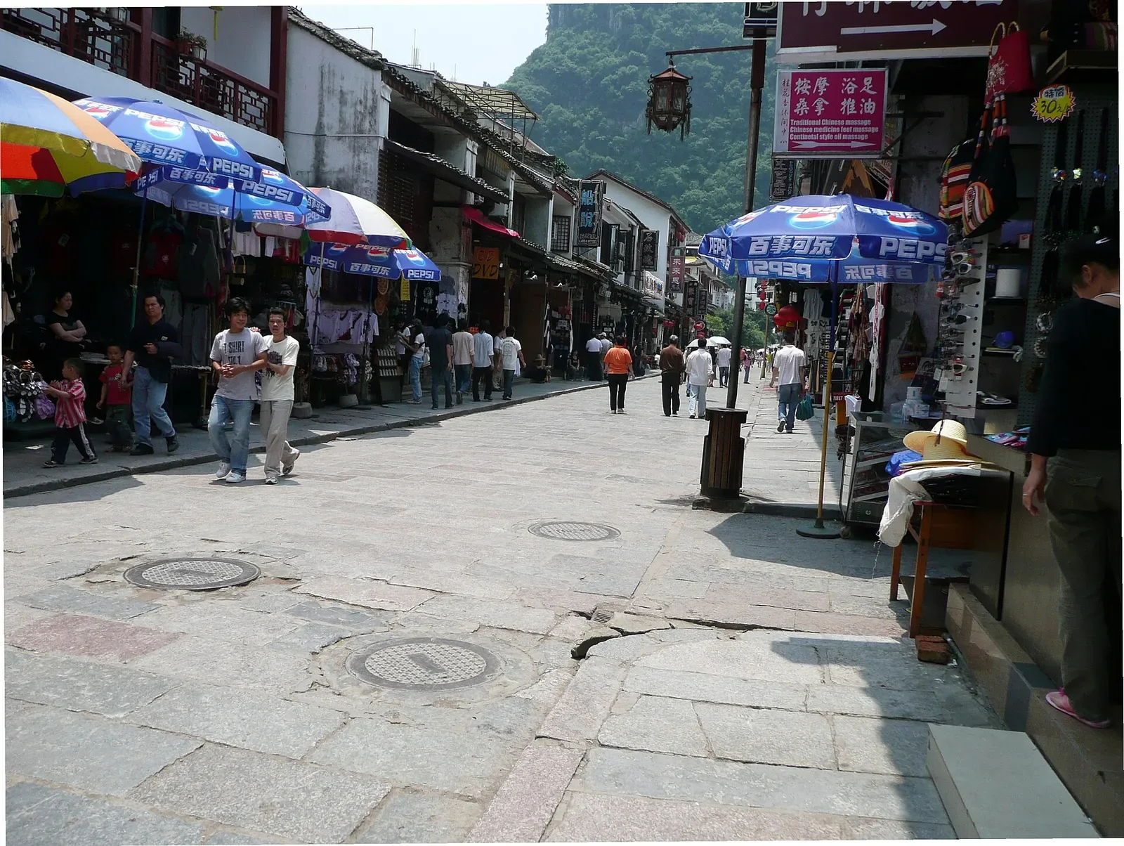 West Street (Xijie) in Yangshuo, Guilin — a 1,400-year-old pedestrian street paved with grey stone slabs, lined with traditional whitewashed shop-houses hung with lanterns and signs, blue Pepsi-branded umbrellas over vendor stalls selling souvenirs, and Chinese and foreign tourists walking and browsing in the late afternoon sun.