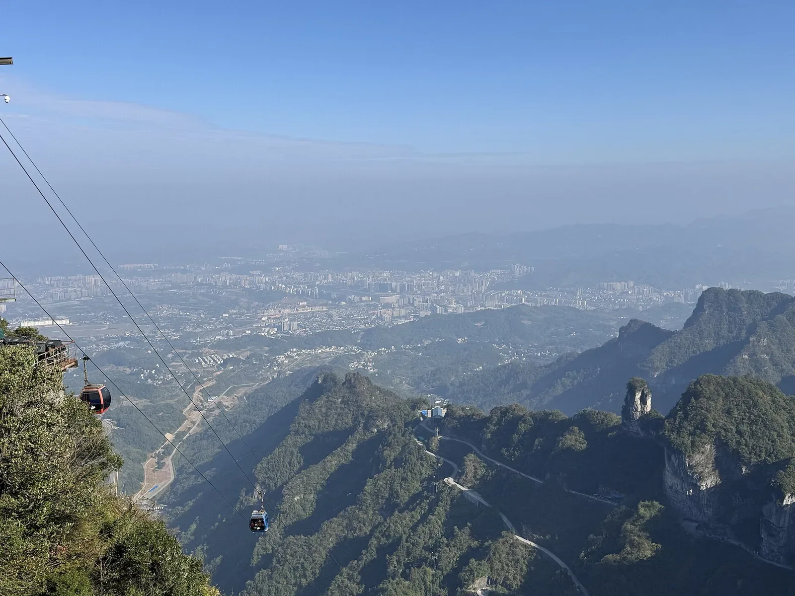 A view from the Tianmen Mountain cable car in Zhangjiajie, looking down at the city and river plain below, with red and blue cable cars climbing up through the sky above forested cliffs and the hazy Cangshan mountain range in the distance.
