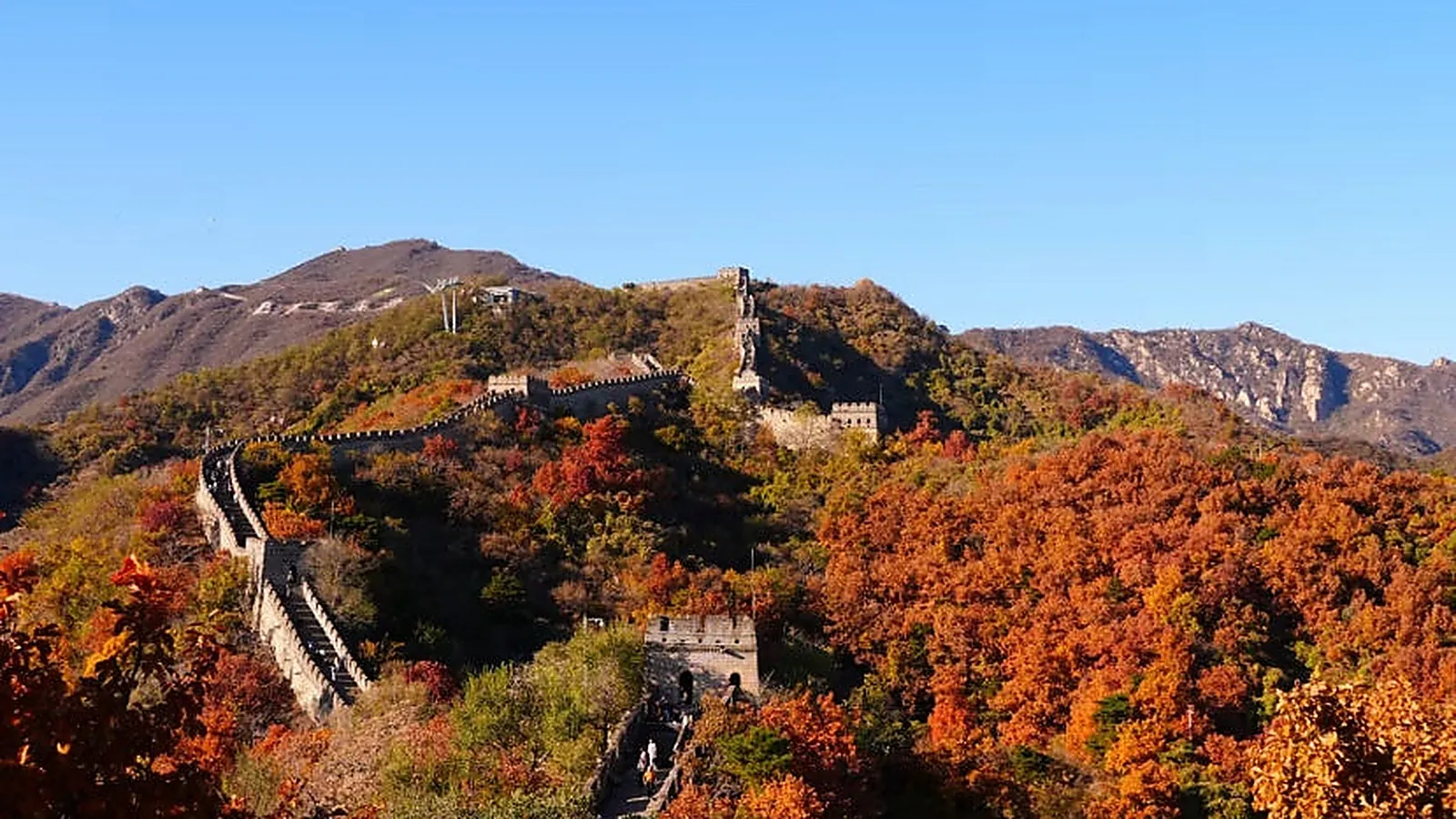 The Great Wall snaking across mountain ridges at sunset, with a Ming-era watchtower silhouetted against an orange-purple sky and autumn foliage glowing in the foreground — the signature late-afternoon light at Mutianyu and Jinshanling that photographers aim for between 4:00 and 5:30 PM