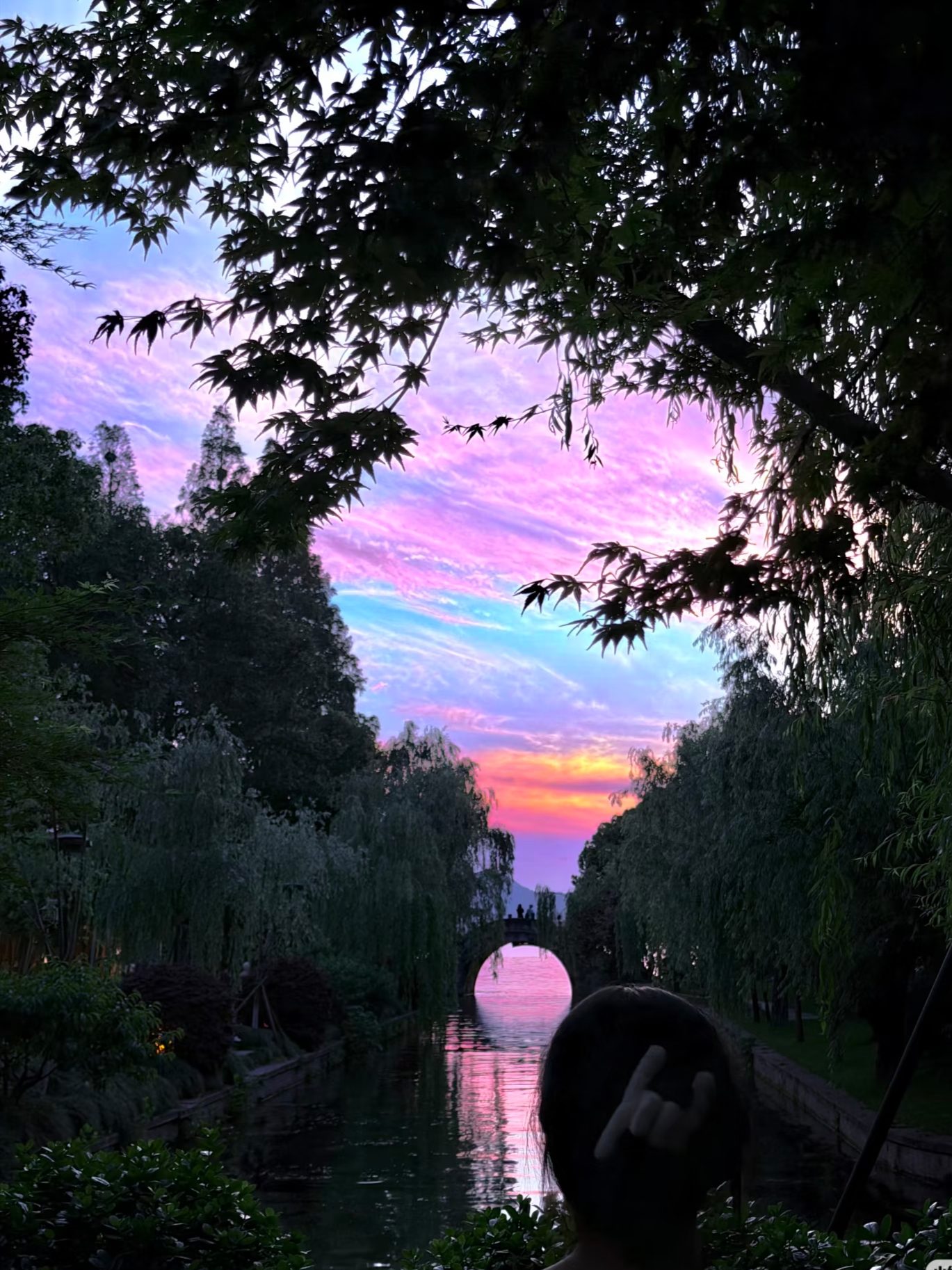 The Leifeng Pagoda silhouetted at sunset behind a single-arch stone bridge over West Lake Hangzhou, with pink and orange sky reflecting in the still water and a few small boats visible in the distance