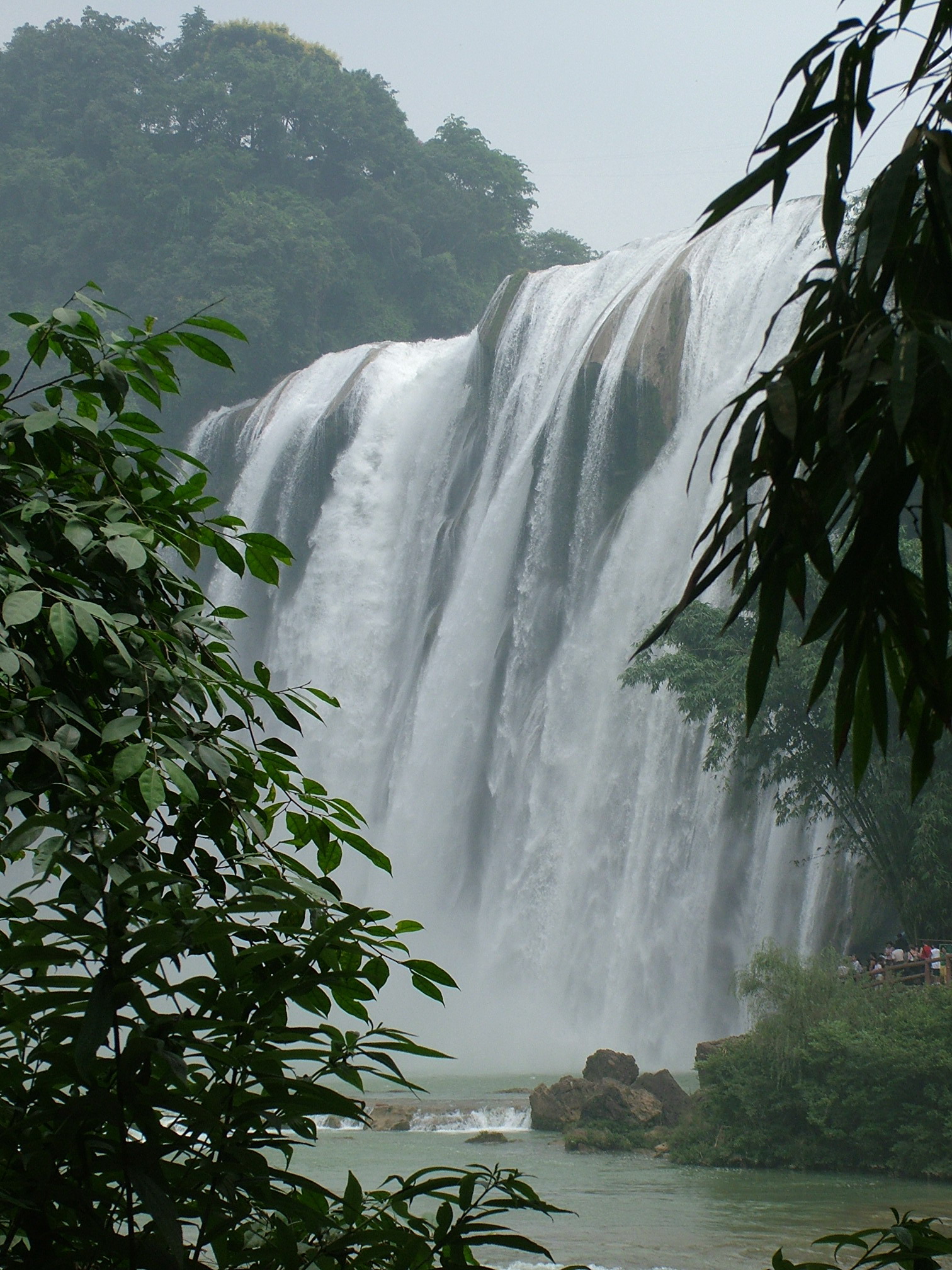 Huangguoshu Waterfall cascading down a wide green cliff in a horseshoe curve, with mist rising from the plunge pool below and lush subtropical forest surrounding the scene, Guizhou, China