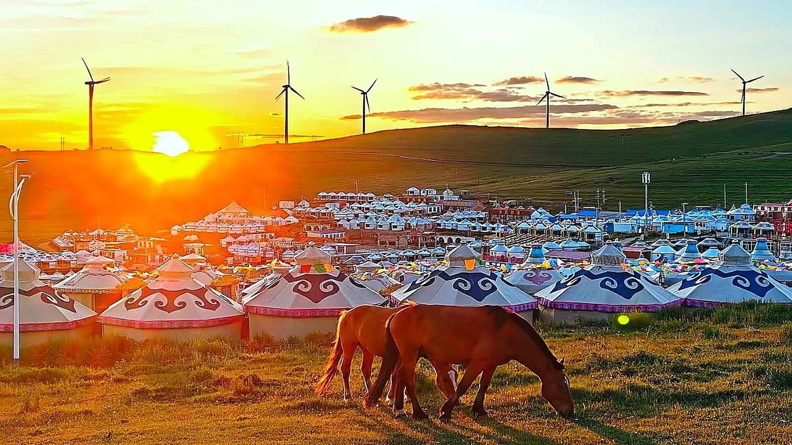 Un tradizionale campo di yurte mongole sulla prateria di Hulunbuir al tramonto: dozzine di gher (yurte) rotonde bianche con bordi blu a motivi nuvolati raggruppati su una bassa collina, un cavallo castano che pascola in primo piano, moderne turbine eoliche sulla linea di cresta sopra e il sole che tramonta in un profondo cielo arancione, la scena classica della steppa di Hulunbuir e lo stesso tipo di campo che il secondo giorno del tour in jeep usa per la sosta notturna, con cavalcate opzionali e cene tradizionali a base di latticini mongoli