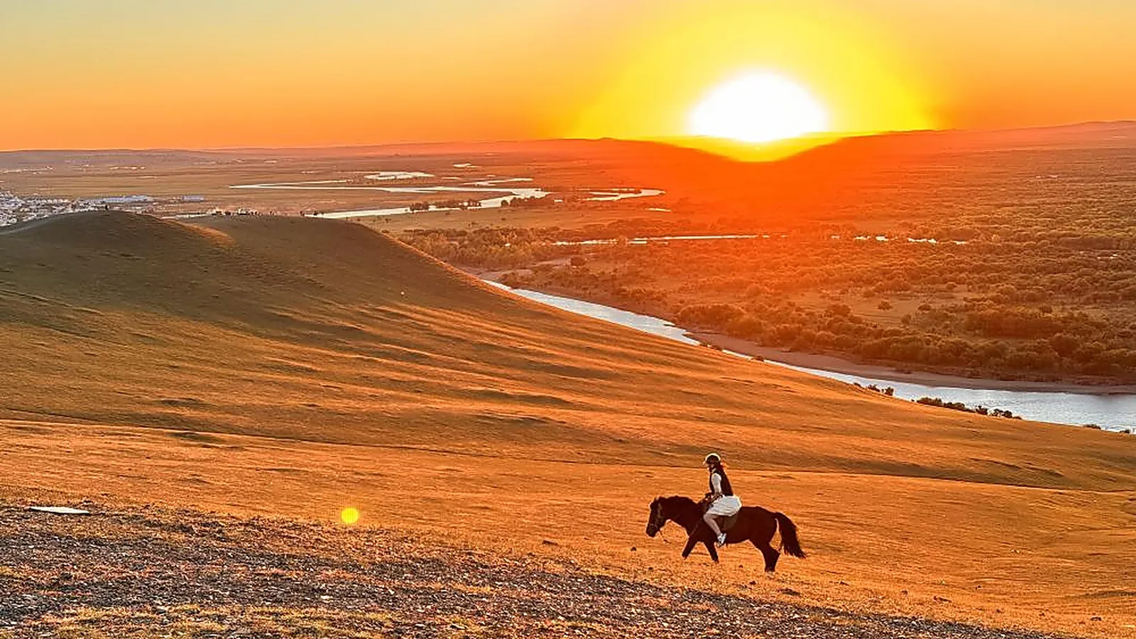 Un cavaliere solitario a cavallo che attraversa le dune di sabbia ondulate di Hulunbuir al tramonto: una sola persona in camicia bianca e casco da equitazione silhouette contro le curve arancioni della duna, il fiume Ergun che serpeggia nella prateria in lontananza, il cielo pieno dell'oro e del rosa della luce serale, l'immagine singola più fotografata di un cavaliere nella steppa di Hulunbuir e il tipo di scatto che la maggior parte dei visitatori stranieri ottiene il secondo giorno del tour in jeep da Enhe a Shiwei