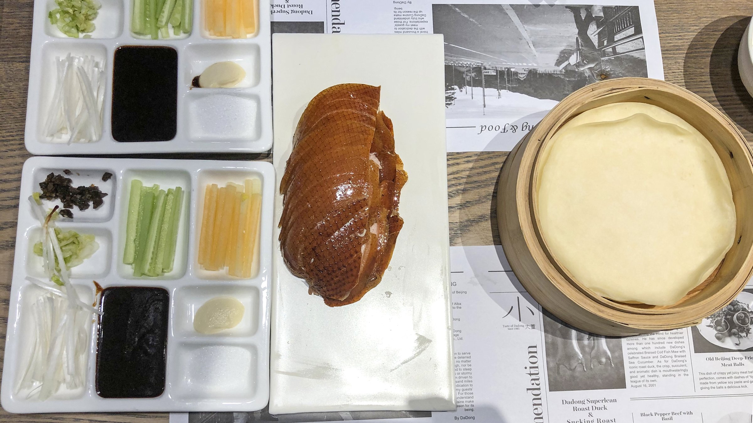 Peking duck guide — whole roasted duck with crispy skin served on a white plate with hoisin sauce, scallions, and thin pancakes in a bamboo steamer