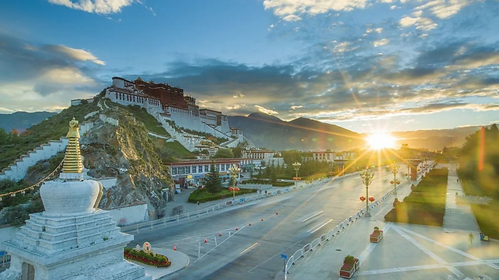 Istana Potala di Lhasa saat matahari terbenam, dilihat dari seberang Beijing Middle Road — seluruh kompleks istana 13 lantai Istana Merah dan Istana Putih yang bertengger di Bukit Merah pada 3.650m, dengan stupa putih sirkuit peziarah Kuil Jokhang di latar depan dan jalan raya kota Lhasa yang lebar dengan lampu jalan menuju ke istana — pemandangan Istana Potala yang paling banyak difoto di panduan perjalanan modern mana pun dan pemandangan yang paling sering difoto pengunjung saat senja pada hari ke-1 perjalanan Tibet mereka