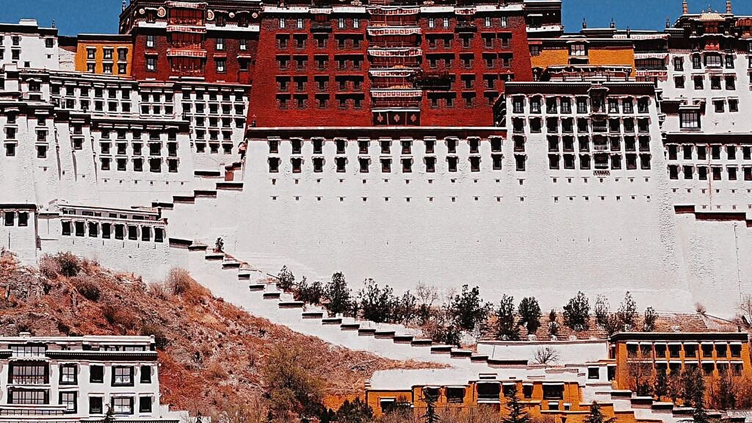 The Potala Palace in Lhasa, Tibet, rising on its red hill with the central Red Palace and flanking White Palace against a clear deep-blue sky, with the surrounding Lhasa valley stretching out below at dawn